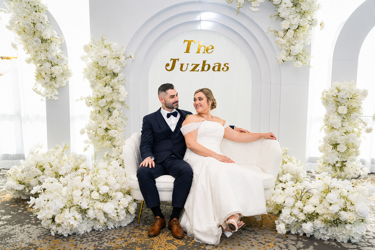 A newlywed couple sitting on a cream-colored sofa, surrounded by white floral decorations, with a backdrop that says 'The Juzbas' in gold letters, in a bright, elegant wedding setting. Chicago events decor photo booth rental