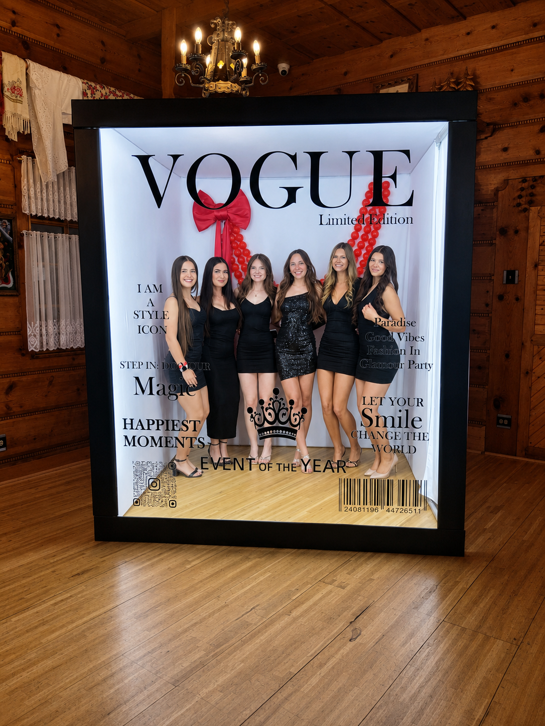 Five women in black dresses standing inside a photo booth with Vogue Limited Edition branding, wooden walls, chandelier, and decorative curtains in the background. Chicago events decor photo booth rental