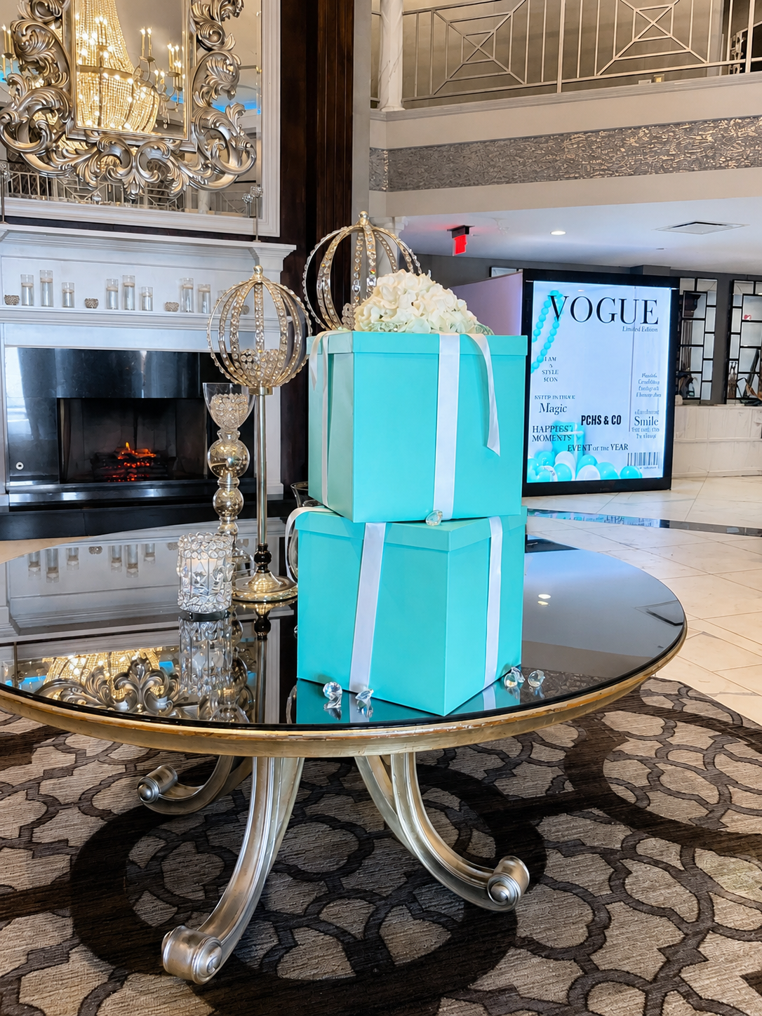 Decorative display with two blue gift boxes with white ribbons, topped with white flowers, on a reflective glass table. Surrounding items include gold and silver crown-shaped ornament, candles, and a mirror. In the background, there is a fireplace, o