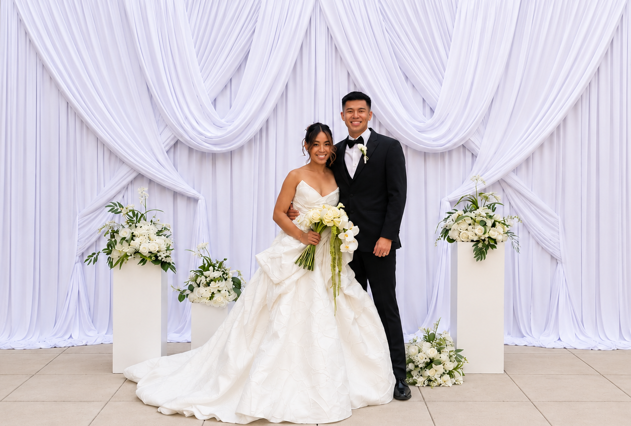 A bride and groom stand together at their wedding, smiling, with white floral arrangements and draped white fabric backdrop. Chicago events decor photo booth rental