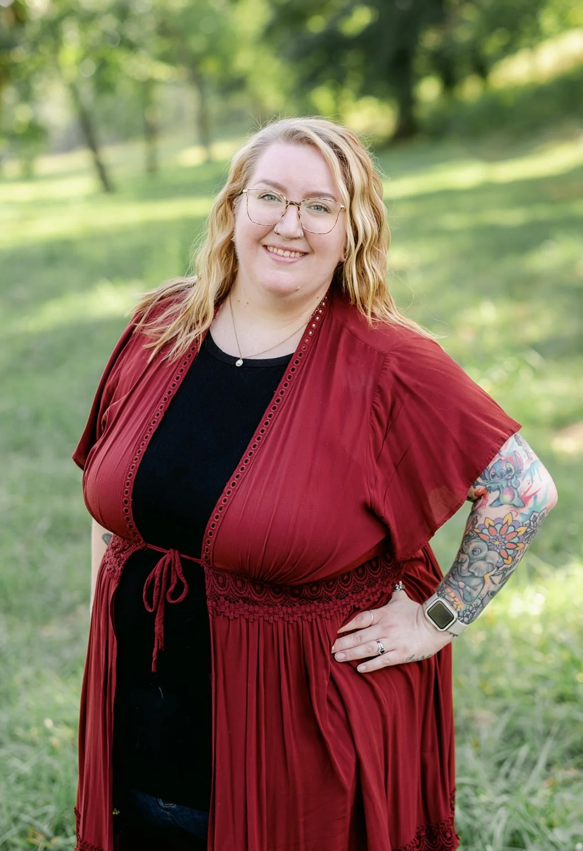 A smiling woman with blonde wavy hair, glasses, and tattoos on her right arm, standing outdoors in a green park with trees, wearing a black top and a red dress with lace details.