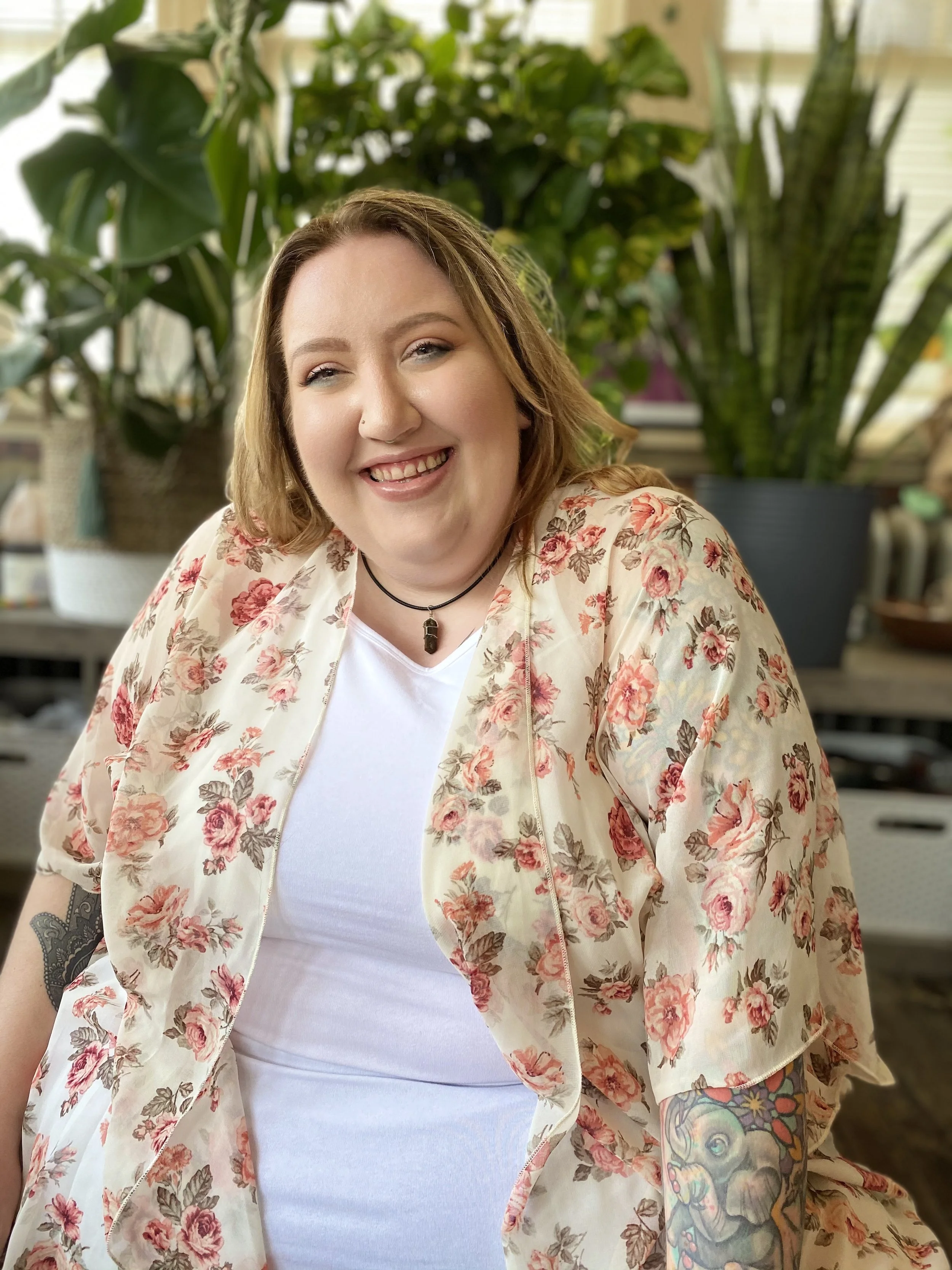 A smiling woman with blonde hair, wearing a floral jacket over a white shirt, sitting in front of green plants indoors.