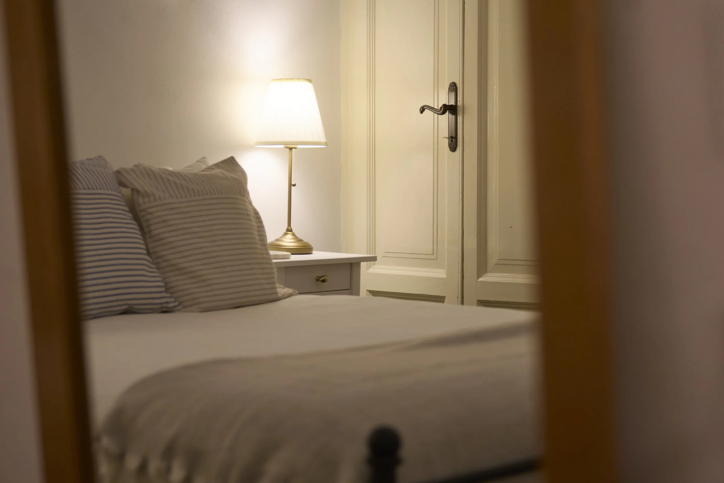 A view of a cozy bedroom with a bed that has striped pillows, a bedside white table, and a warm lamp, seen through a partially opened door.