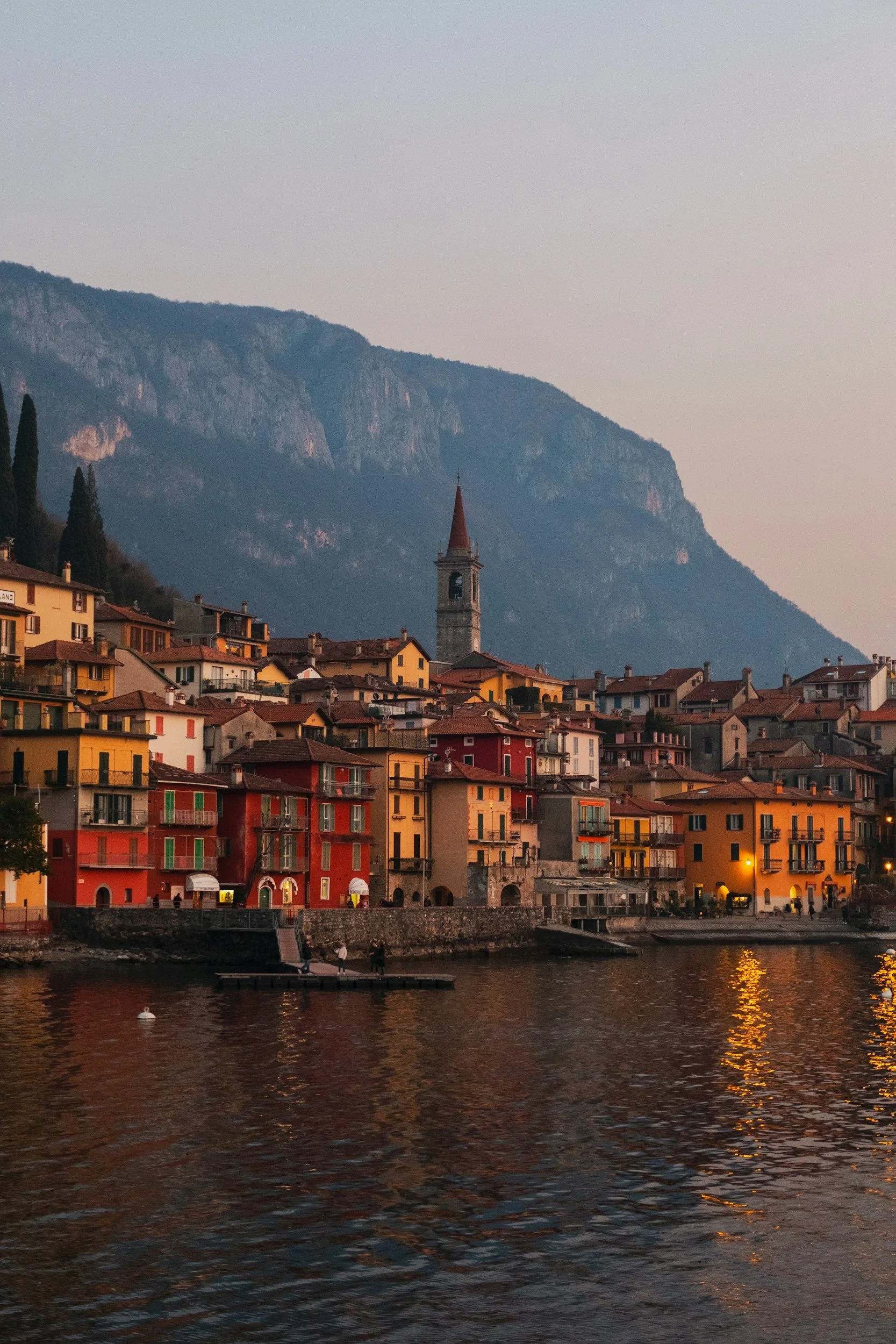 A scenic view of a lakeside town at dusk with colorful buildings, a church with a tall steeple, and mountains in the background. The water reflects the lights from the buildings.