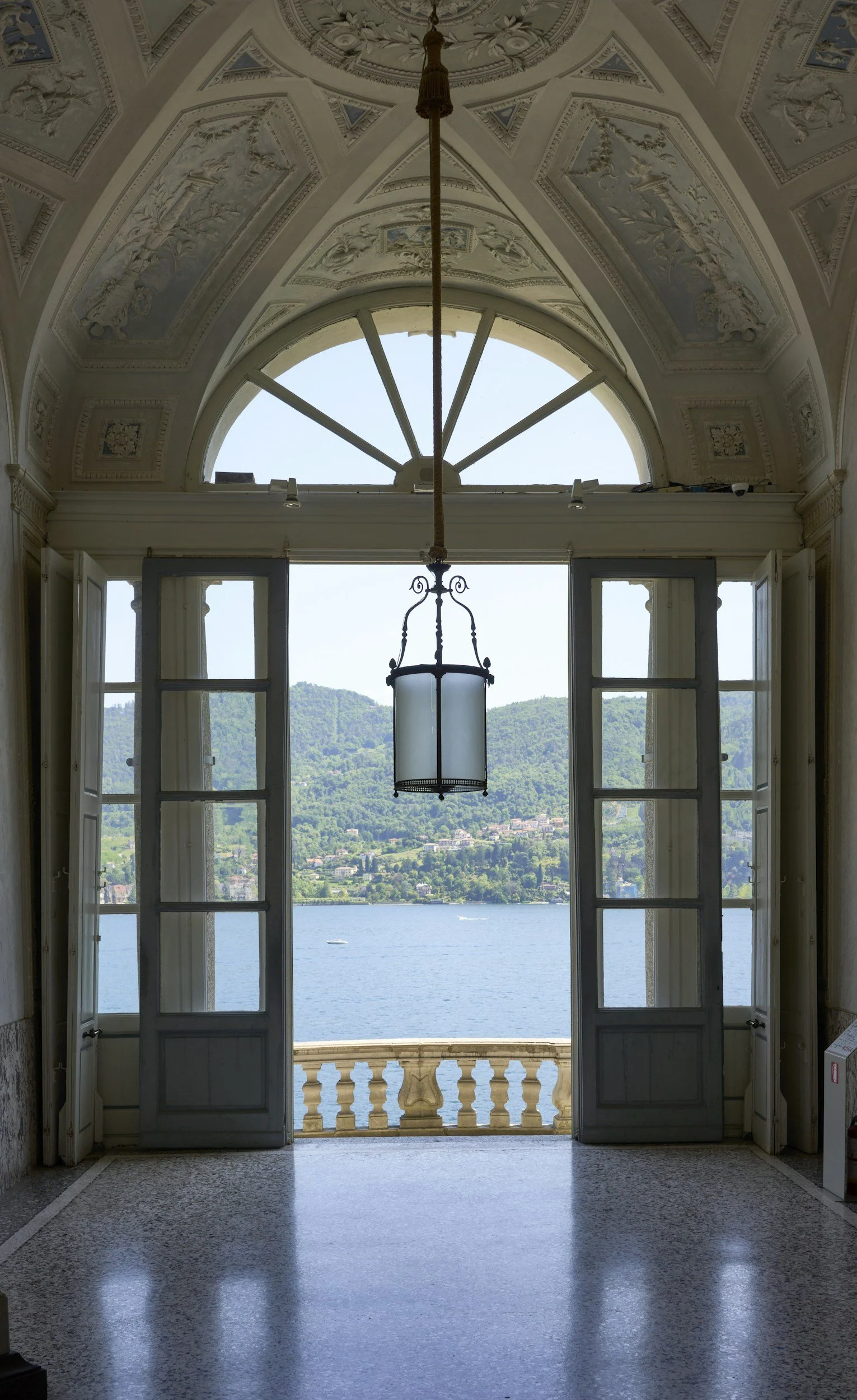 View through open French doors overlooking a lake with green hills and houses in the background, framed by a decorative ornate ceiling and a hanging light fixture.