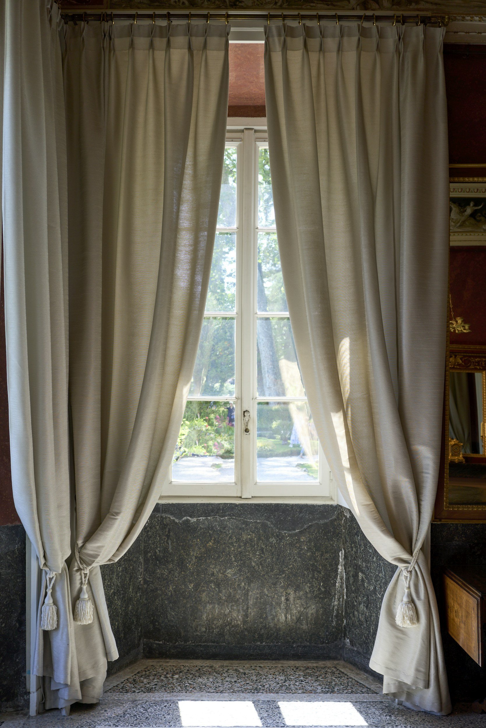 A window with white framing and multiple panes, framed by cream-colored curtains tied back on both sides, revealing a view of a sunny garden with trees and greenery outside.