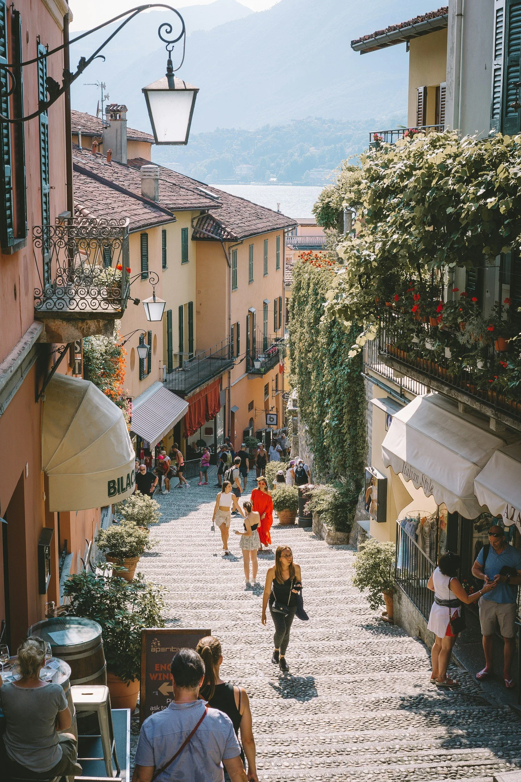 A street scene in a European town with colorful buildings, cobblestone pavement, and people walking and shopping, with a water body and mountains in the background.