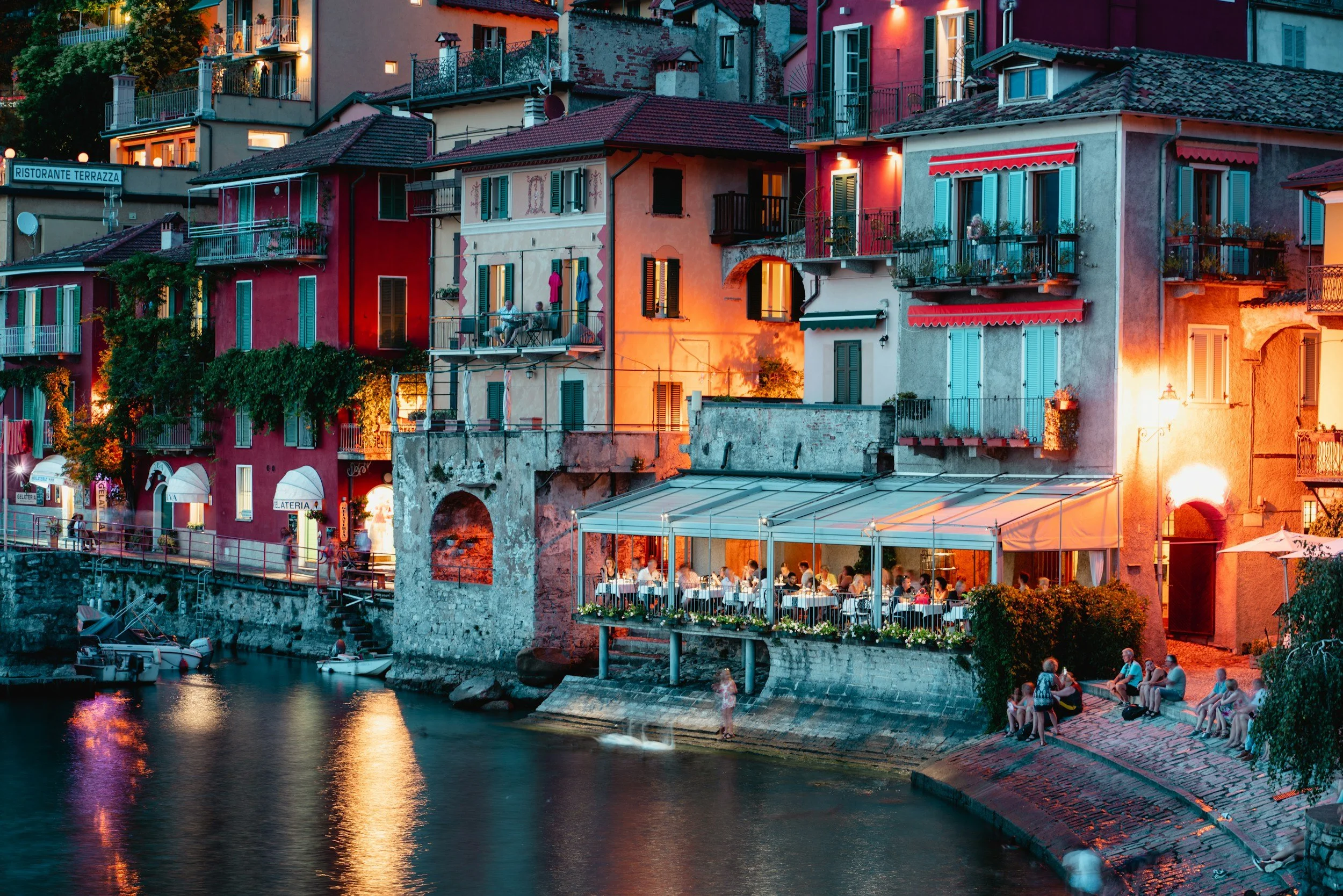 Colorful buildings with balconies and open windows along a waterfront, with people sitting at outdoor tables and walking nearby during evening time.