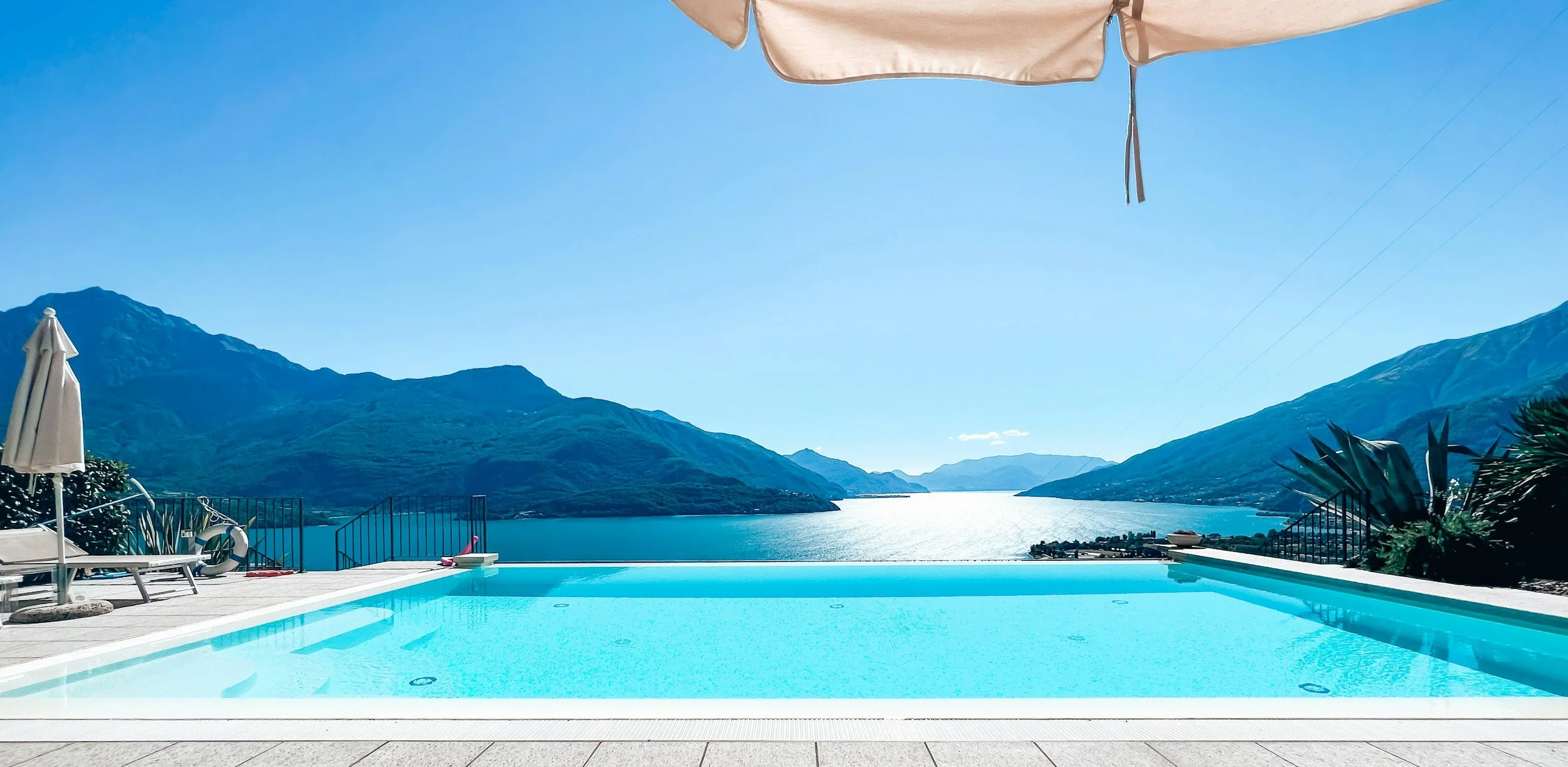 Piscina con vista sul lago e le montagne sotto un cielo chiaro, con ombrelloni e lettini sul bordo piscina.