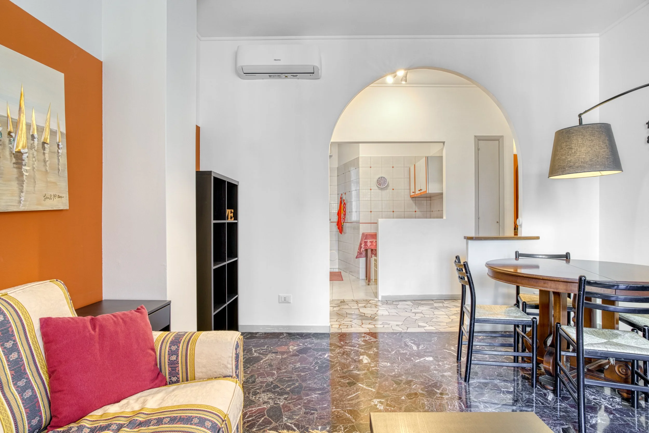 Living room with a beige couch with colorful pillows, a black bookshelf, a dining table with black chairs, a gray lampshade, and an archway leading to a kitchen with tile walls and a red towel hanging.