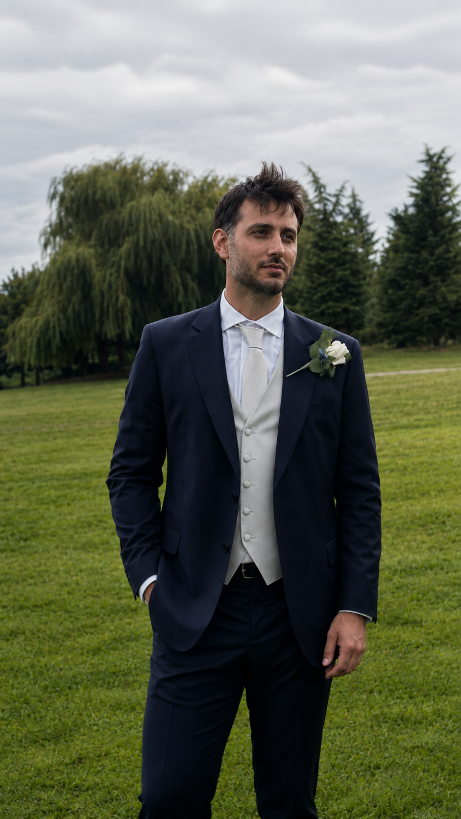 Uomo elegante con abito da sposo blu e cravatta bianca, con fiore sul taschino, in un giardino verde in una giornata nuvolosa.