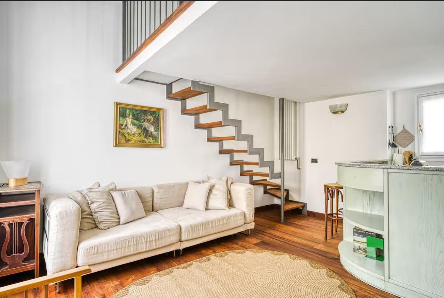 Soggiorno con divano beige, quadro in cornice dorata, scala aperta con gradini di legno e struttura metallica, parete bianca, tavolo da bar verde chiaro, poltrona in legno, parquet in legno, tappeto rotondo.