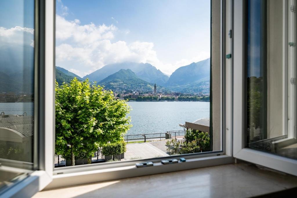 Vista di un lago con montagne sullo sfondo e un cielo sereno, vista da una finestra aperta in una stanza.