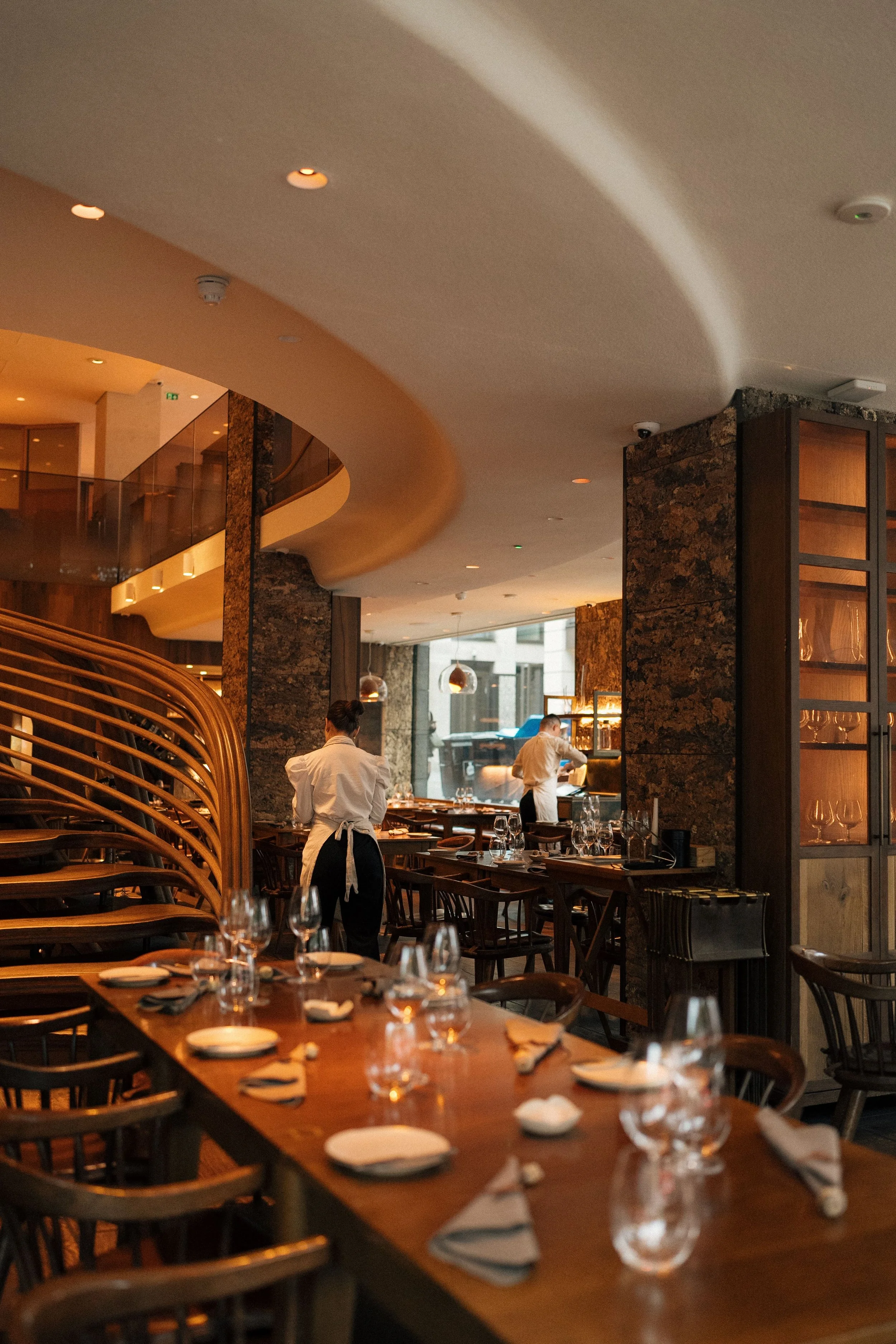 Interior of a restaurant with a warm ambiance, set with wine glasses and plates on wooden tables, with staff preparing and arranging tables, large windows letting in natural light, and decorative stone walls.