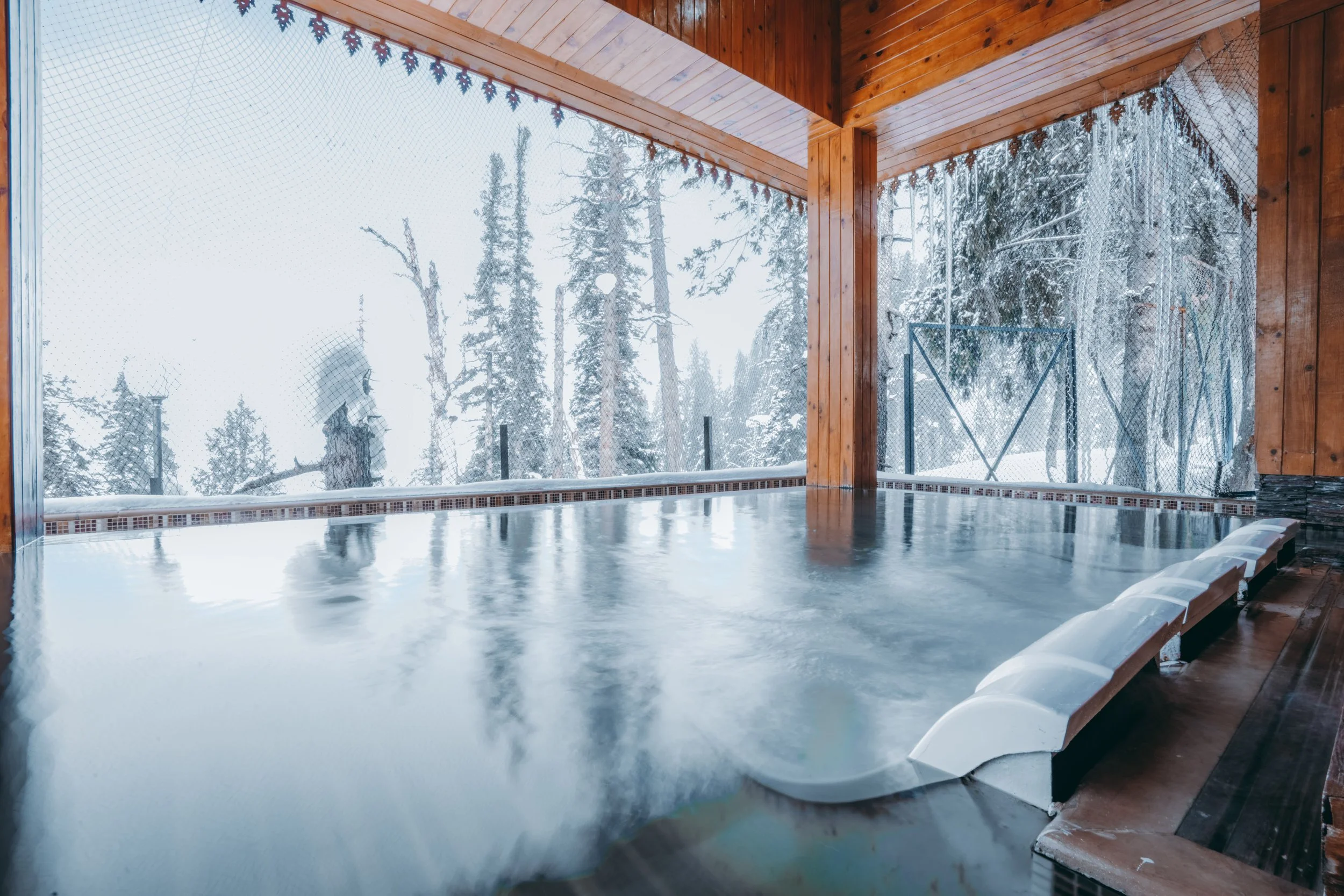 Indoor hot tub with steam and a wood-paneled interior, overlooking a snowy forest through large windows.