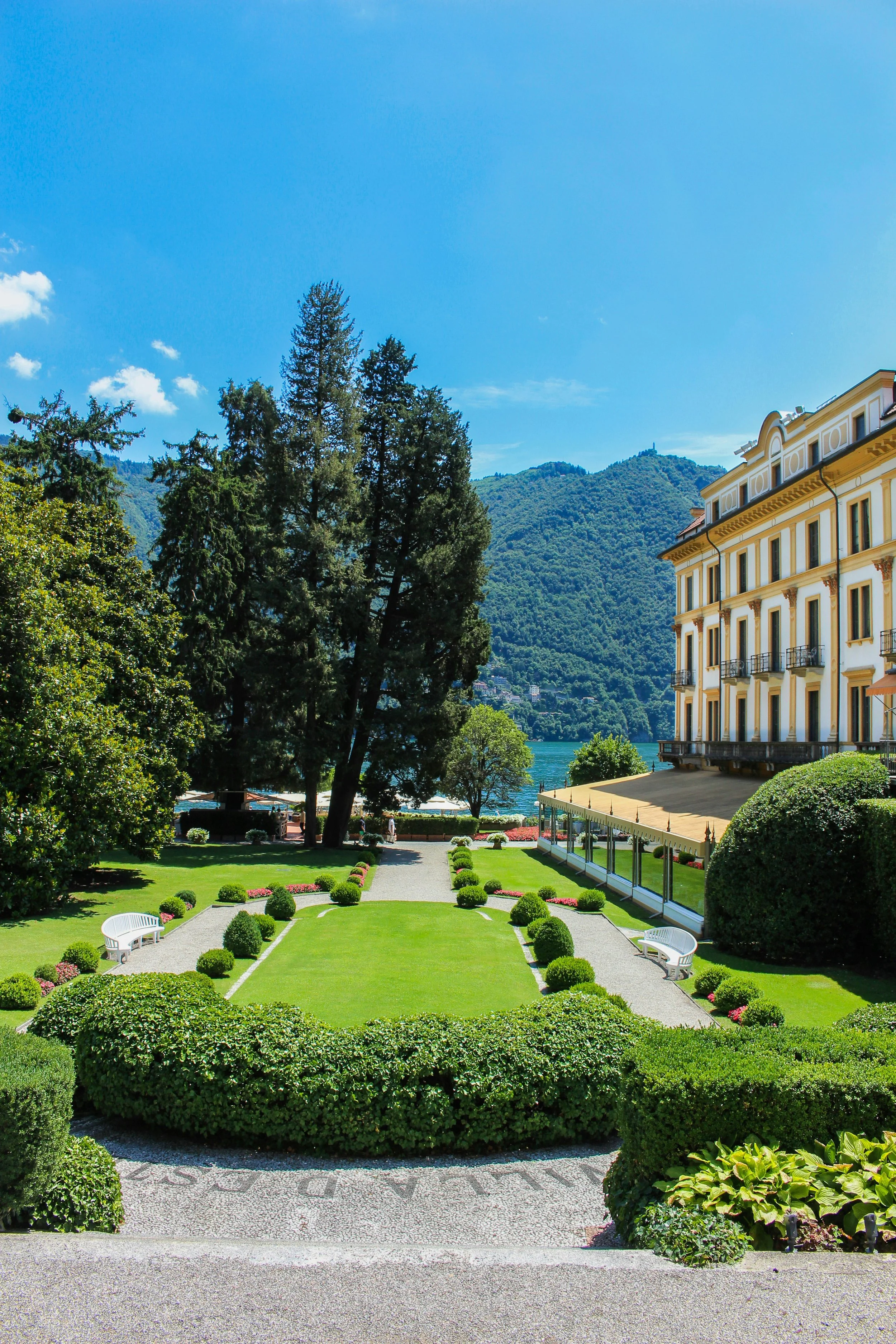 Beautiful landscaped garden with trimmed bushes and benches, overlooking a lake with trees and mountains in the background, sunny day with clear blue sky.