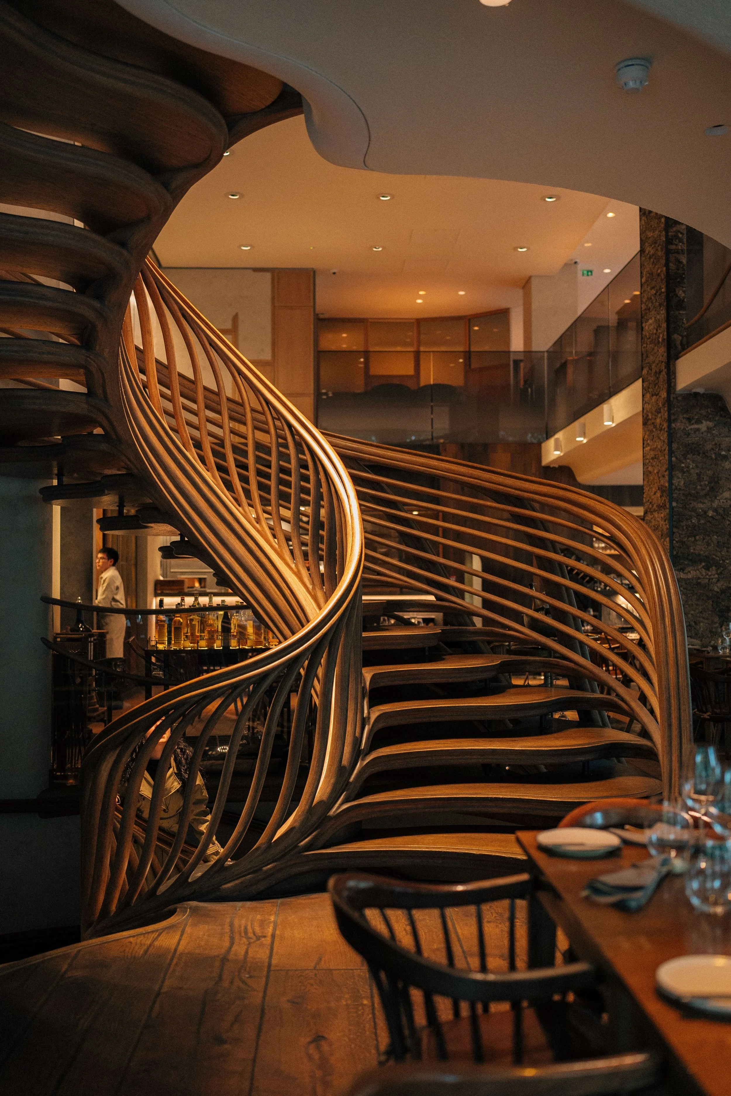 Interior of a restaurant or bar with a wooden railing, tables set with napkins and silverware, and warm lighting.