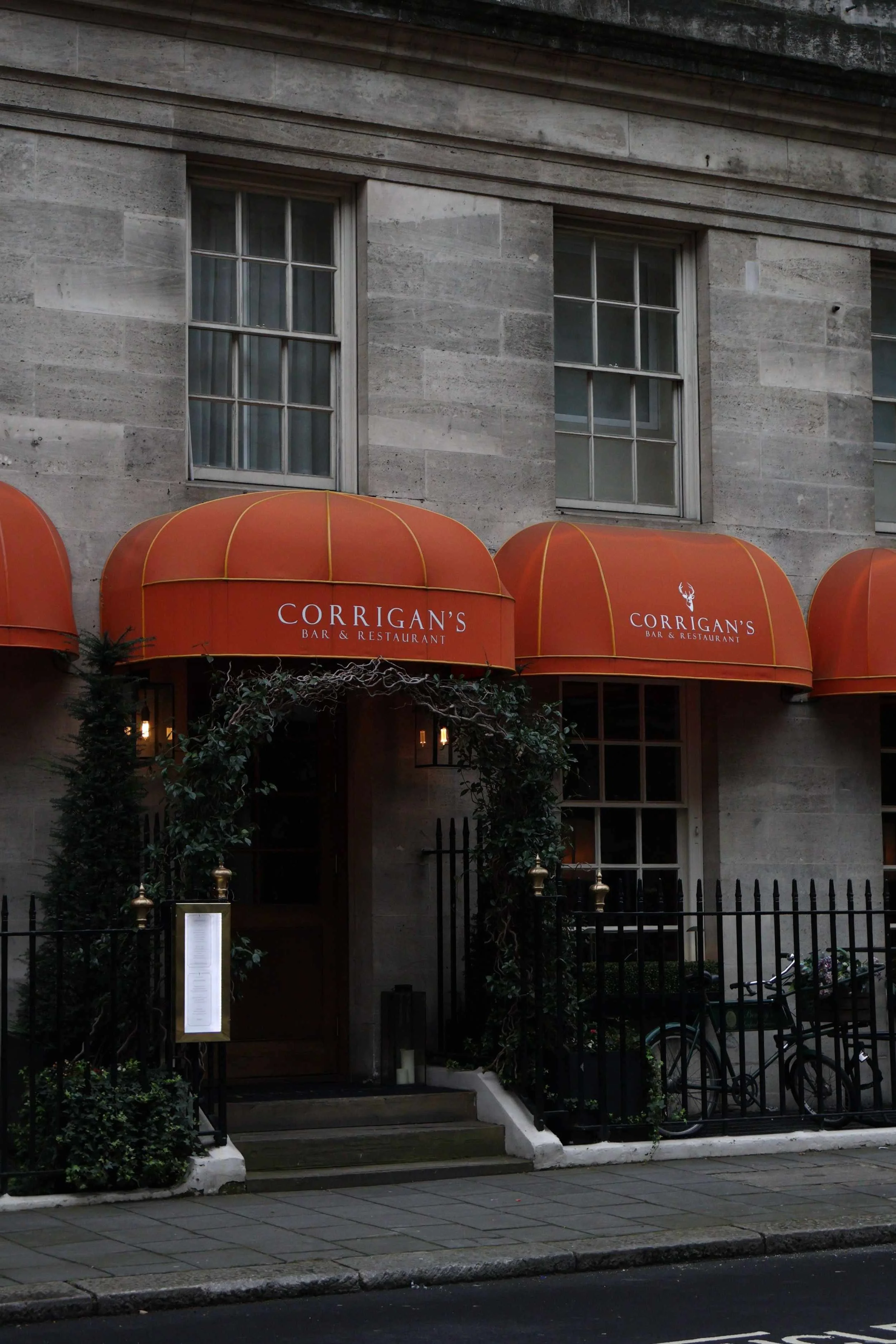 The image shows the entrance to a bar and restaurant called Corrigan's, with a red canopy featuring the establishment's name and logo. The entrance is surrounded by greenery, including small trees and plants, and is fenced with black metal fencing. There are two large windows above the entrance in a stone building facade.