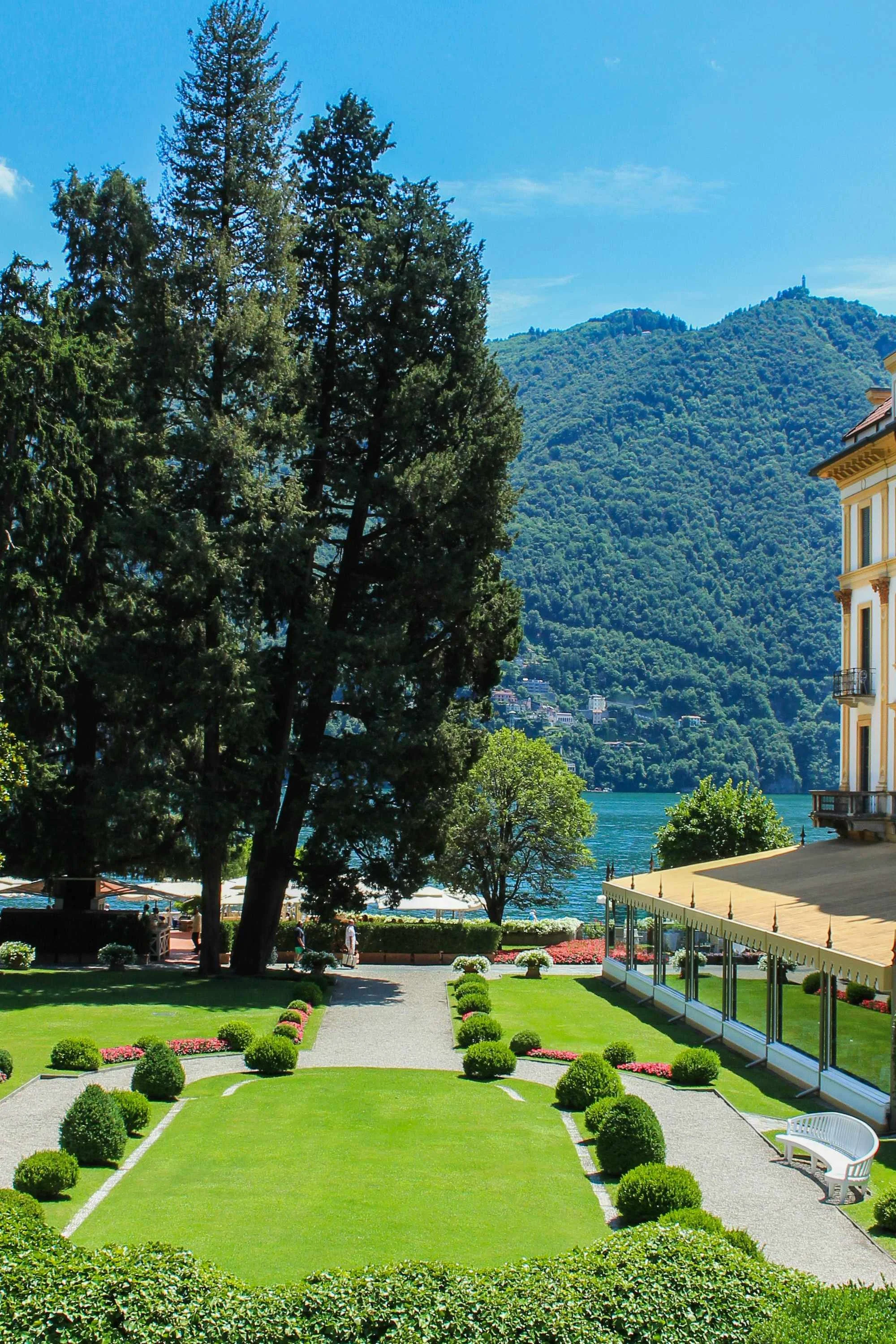 A landscaped garden with a green lawn, small bushes, and flowers, with a pathway, benches, and a building with a yellow roof and white walls. There are large trees, a lake, and hills with buildings in the background, under a blue sky with some clouds.