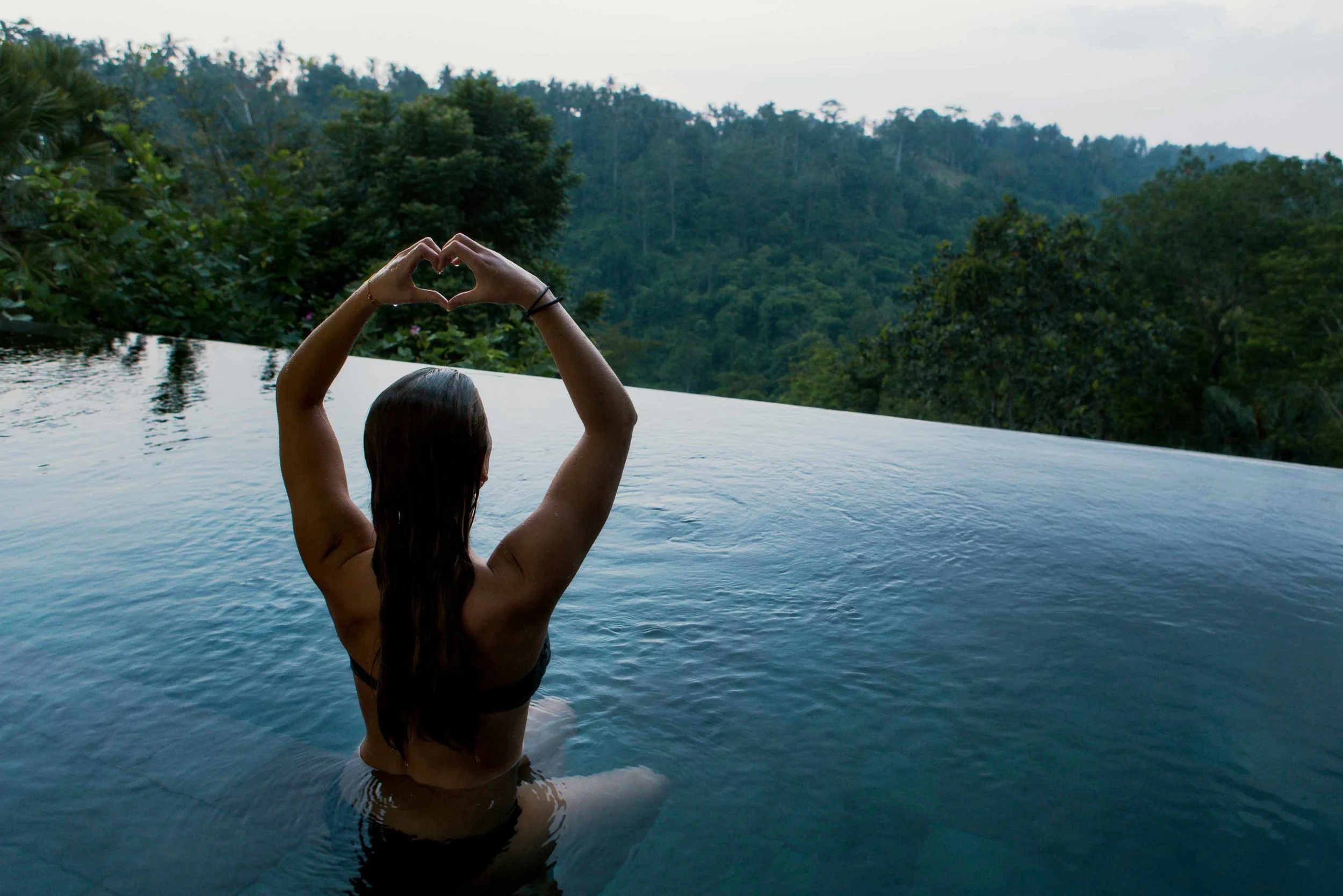 A woman standing in a pool, making a heart shape with her hands, with a background of lush green trees and a cloudy sky in a natural outdoor setting.