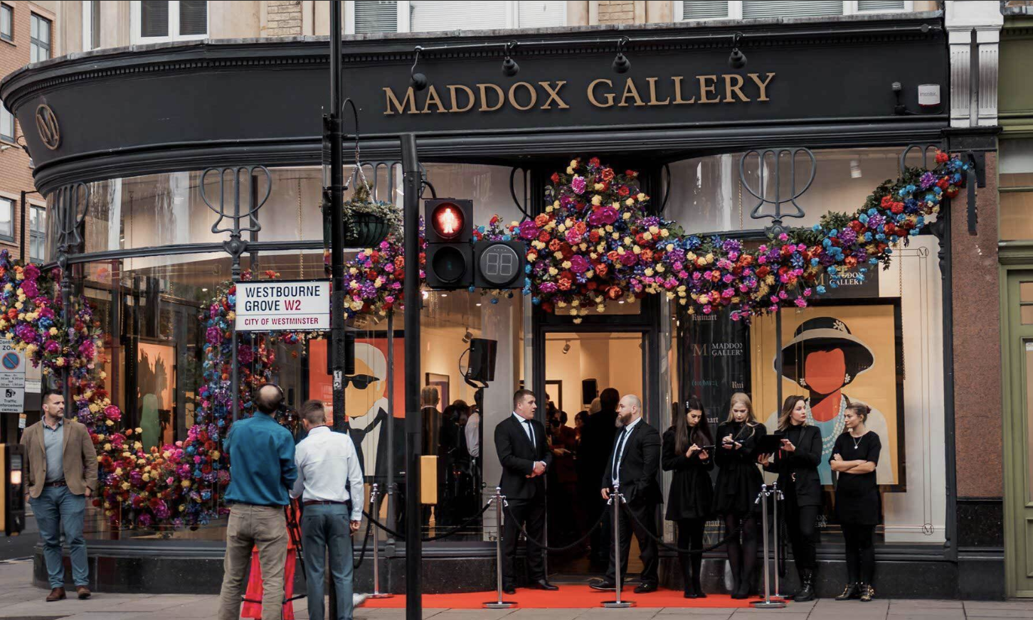People gathered outside Maddox Gallery, a contemporary art gallery in Westminster, London, decorated with colorful flowers and artwork visible through the large glass windows.