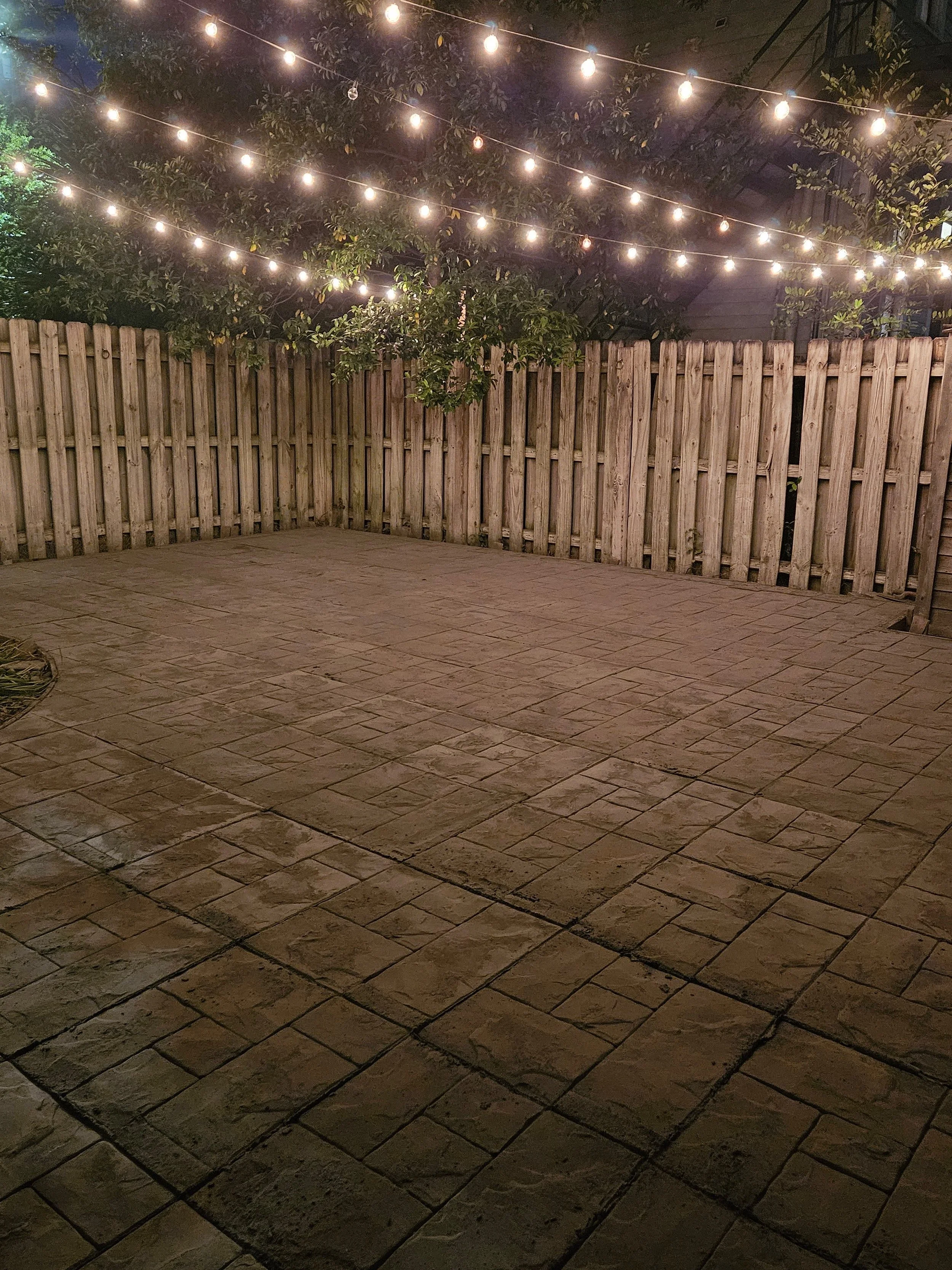 Nighttime backyard patio with stone pavers, wooden fence, trees, and string lights hanging overhead.