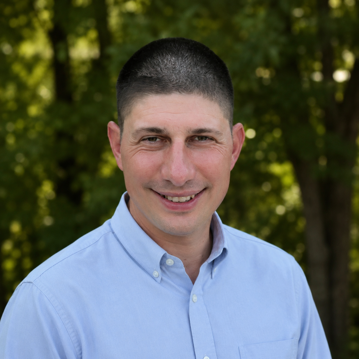 A smiling man with short dark hair and light skin, wearing a light blue button-up shirt, outdoors with green trees in the background.