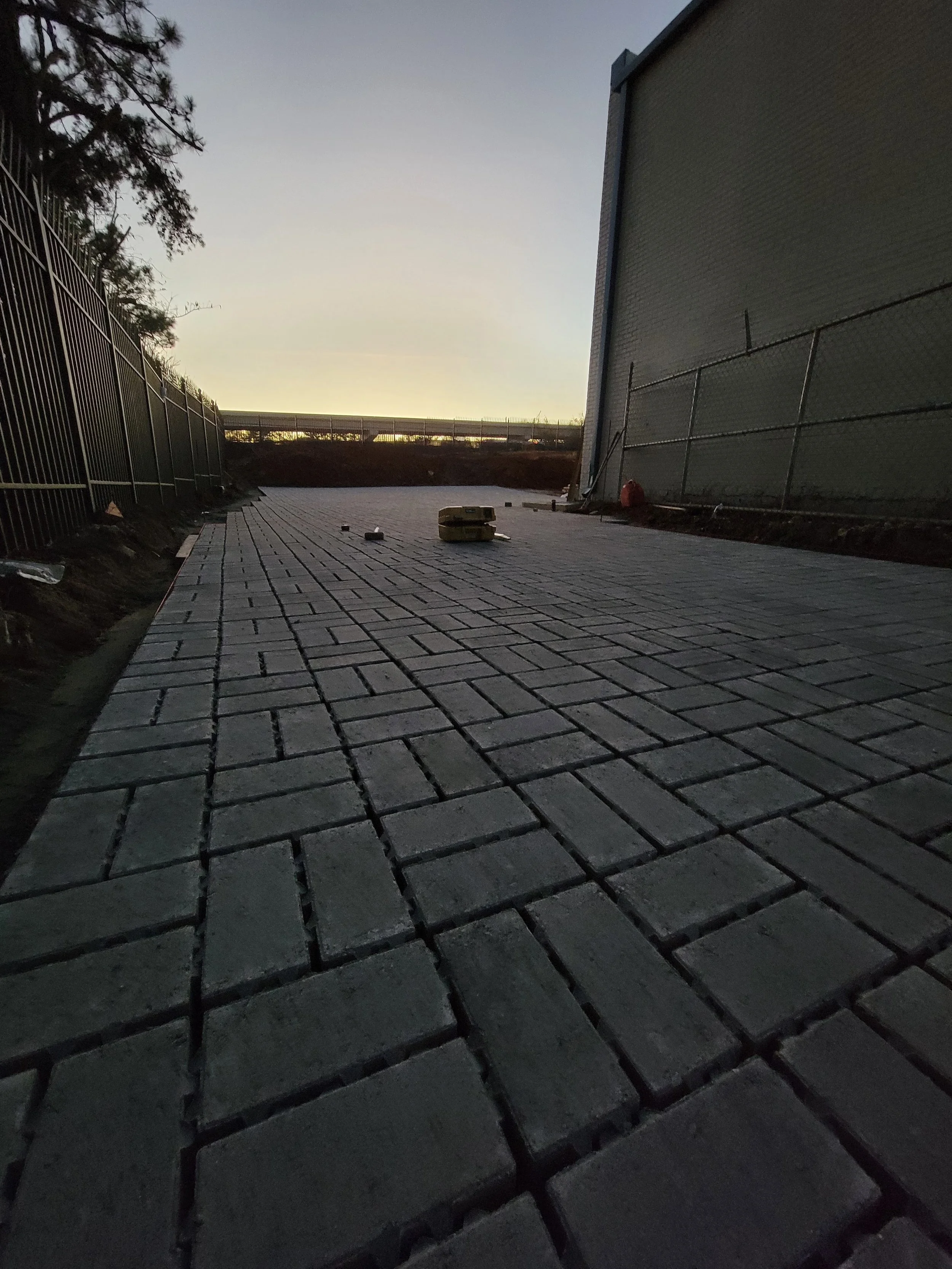 A paved walkway under construction with rectangular bricks laid out in progress, surrounded by fencing on both sides, at dusk with a faint sunset in the background.