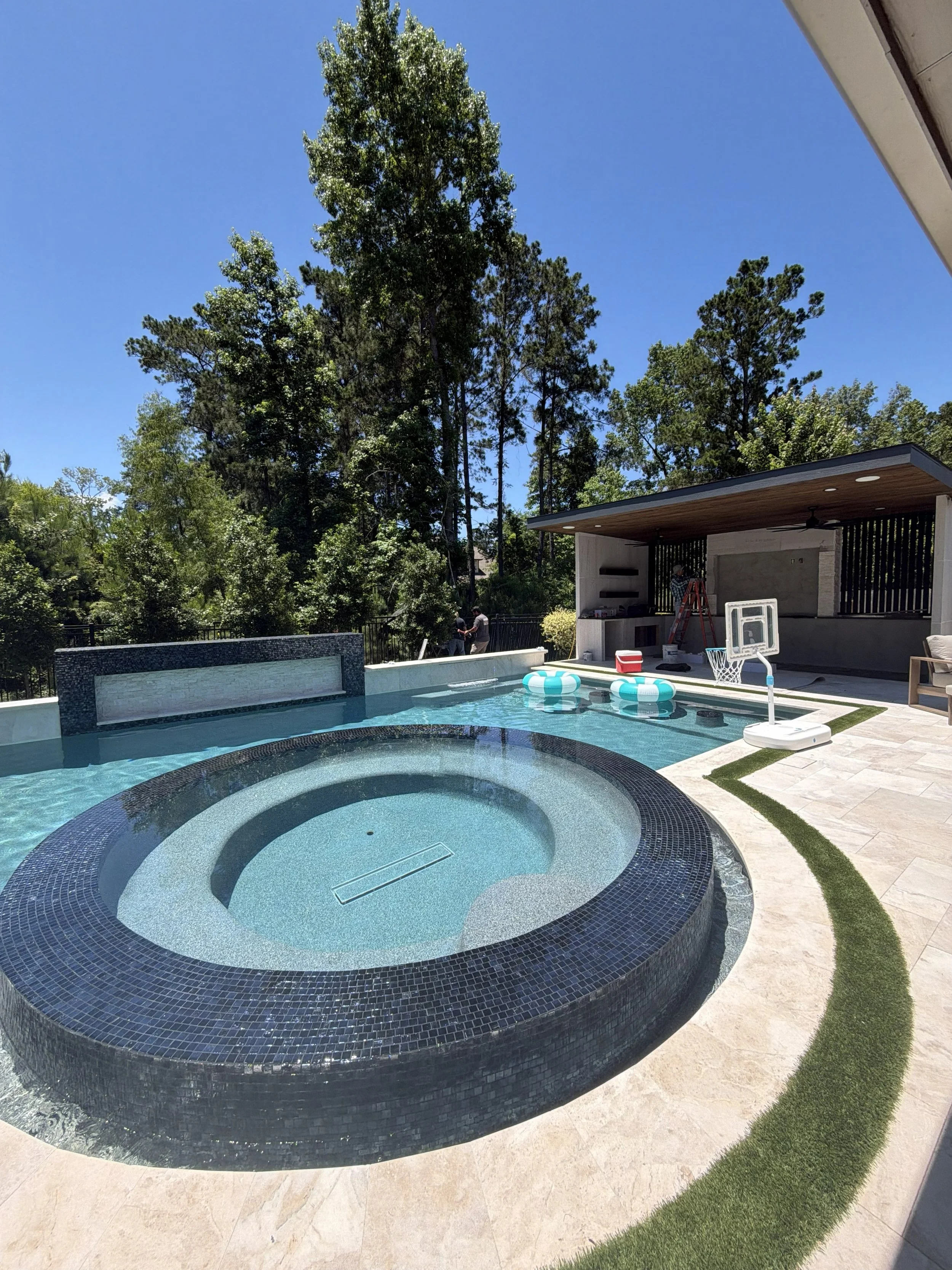 A swimming pool with a built-in hot tub, surrounded by a tiled deck, with a modern outdoor structure and tall trees in the background. There are inflatable pool floats in the water and some people working on the setup.