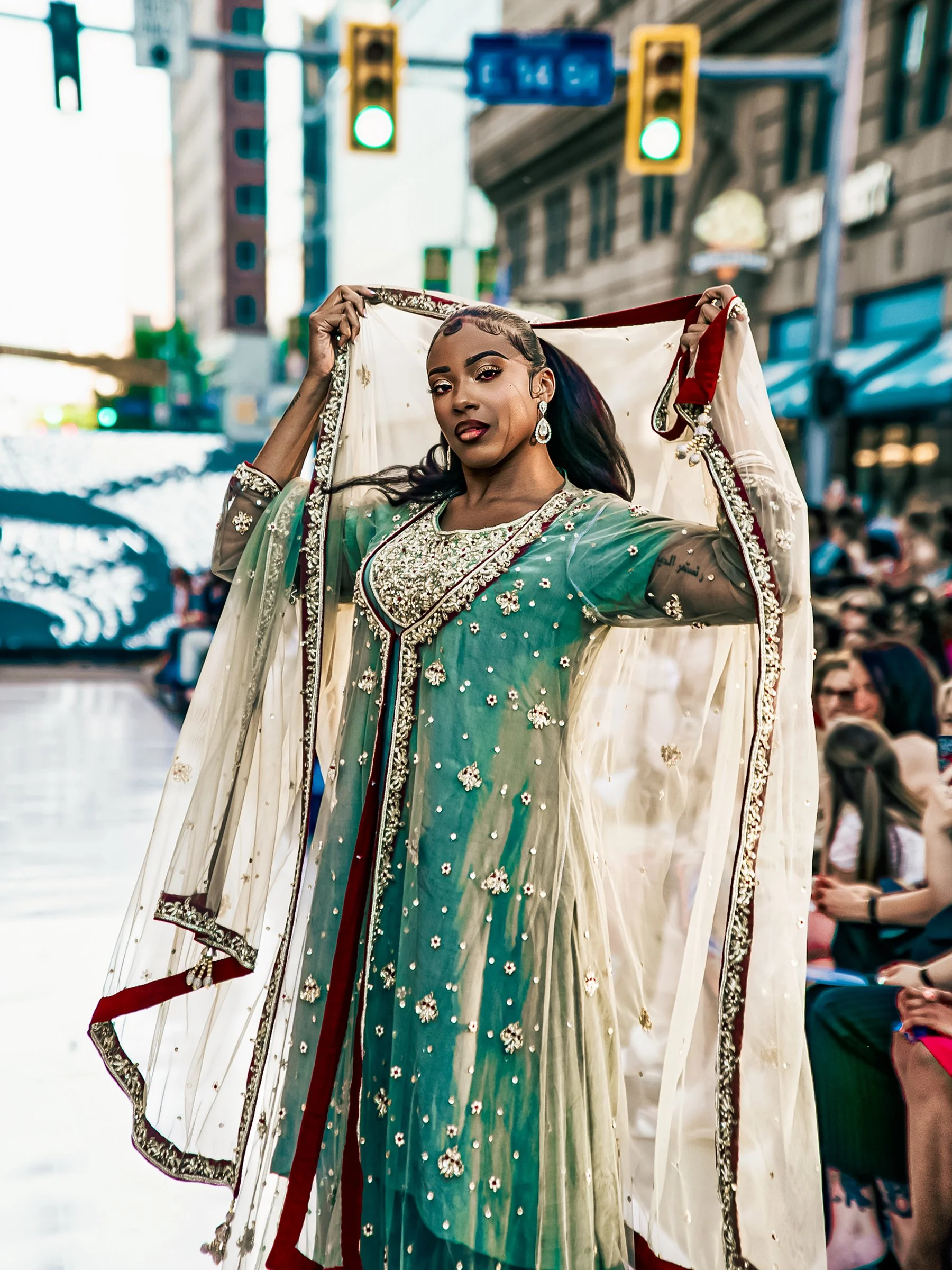 A woman wearing a traditional South Asian dress with detailed embroidery, standing outdoors in an urban setting with traffic lights and people in the background.