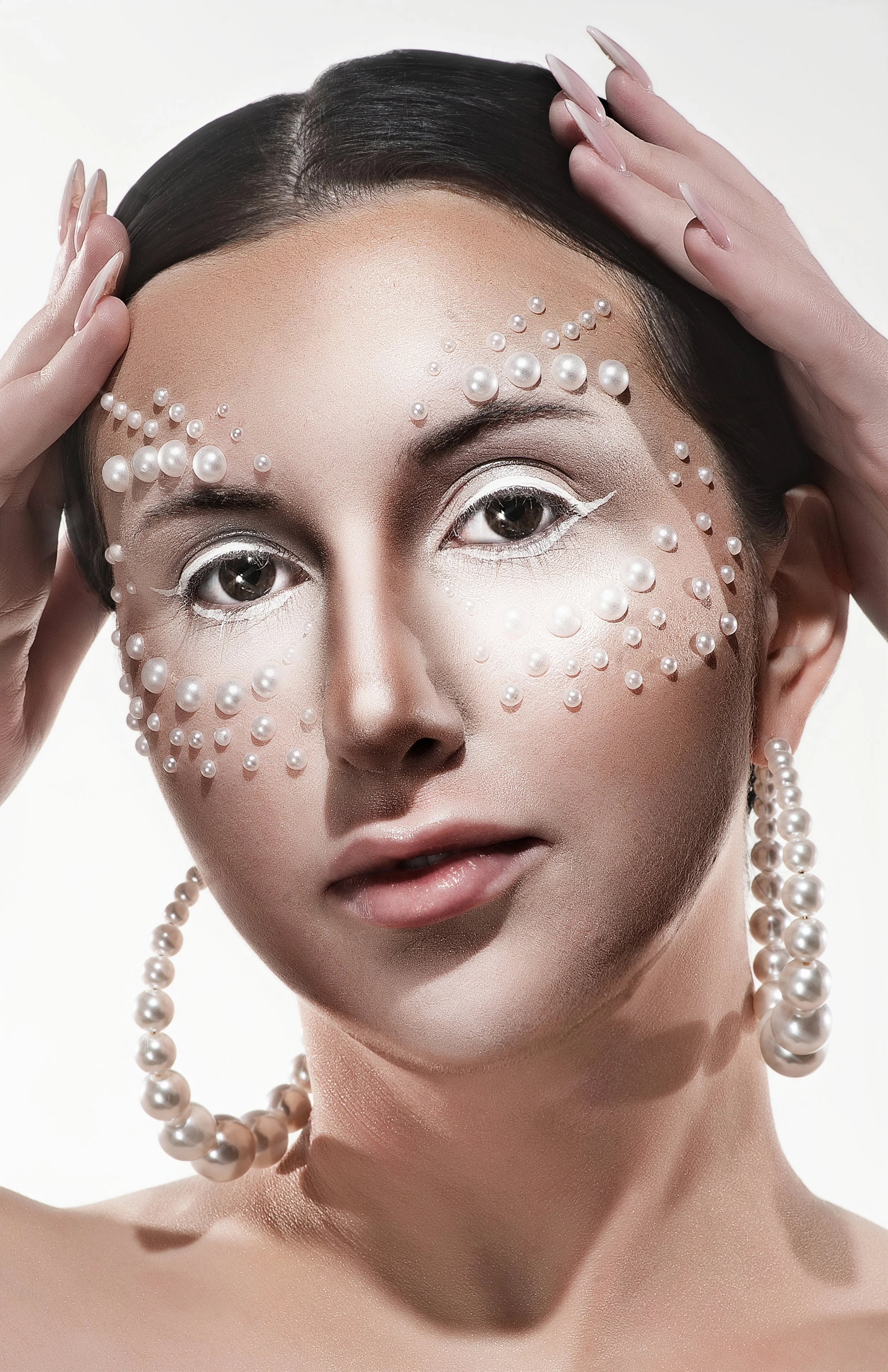 A close-up portrait of a woman with pearl embellishments on her face, slicked-back dark hair, and pearl jewelry. She holds her hands near her head, with manicured nails, against a plain white background.