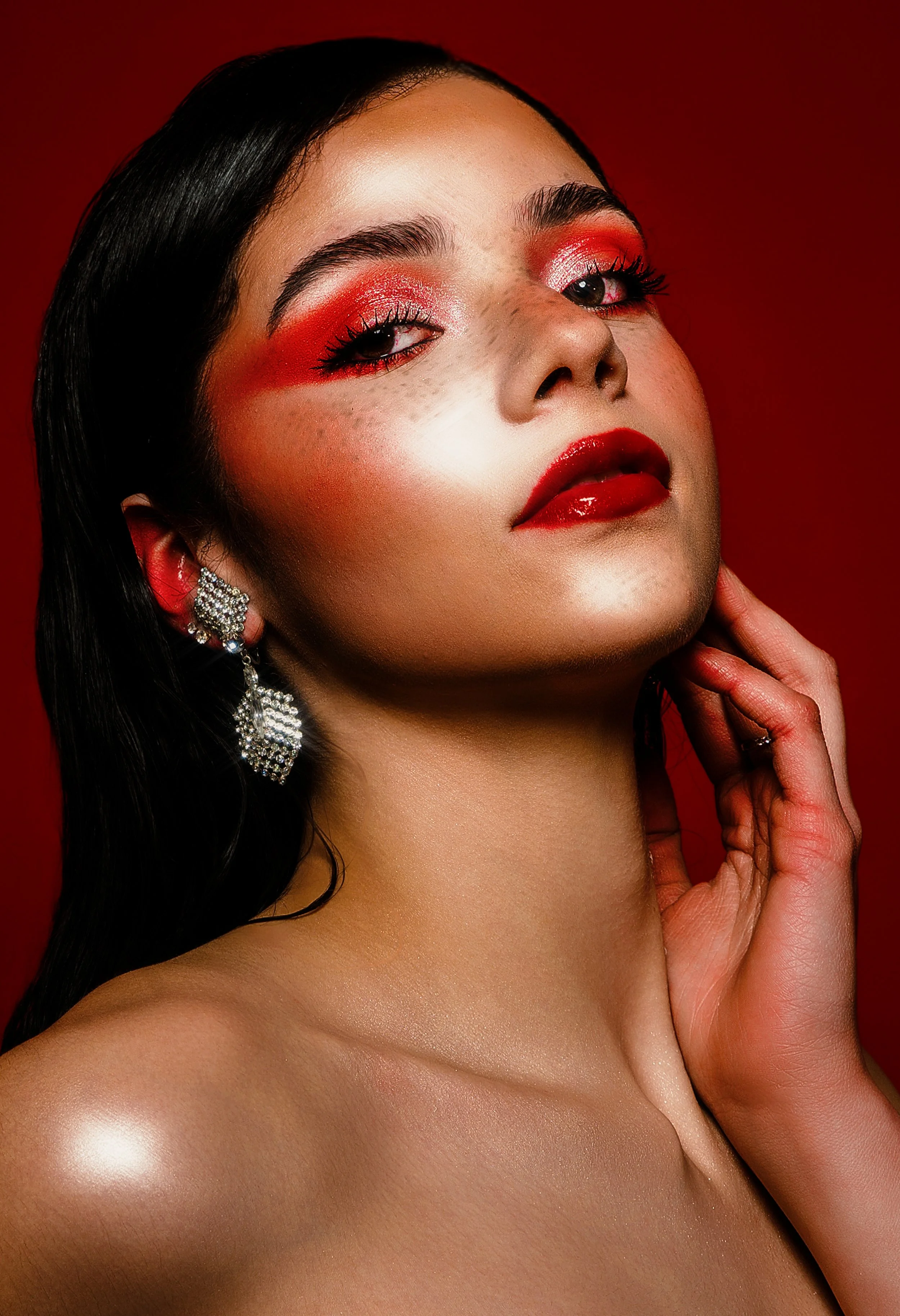 A woman with long dark hair, wearing statement earrings, with bright red makeup, and posing against a red background.