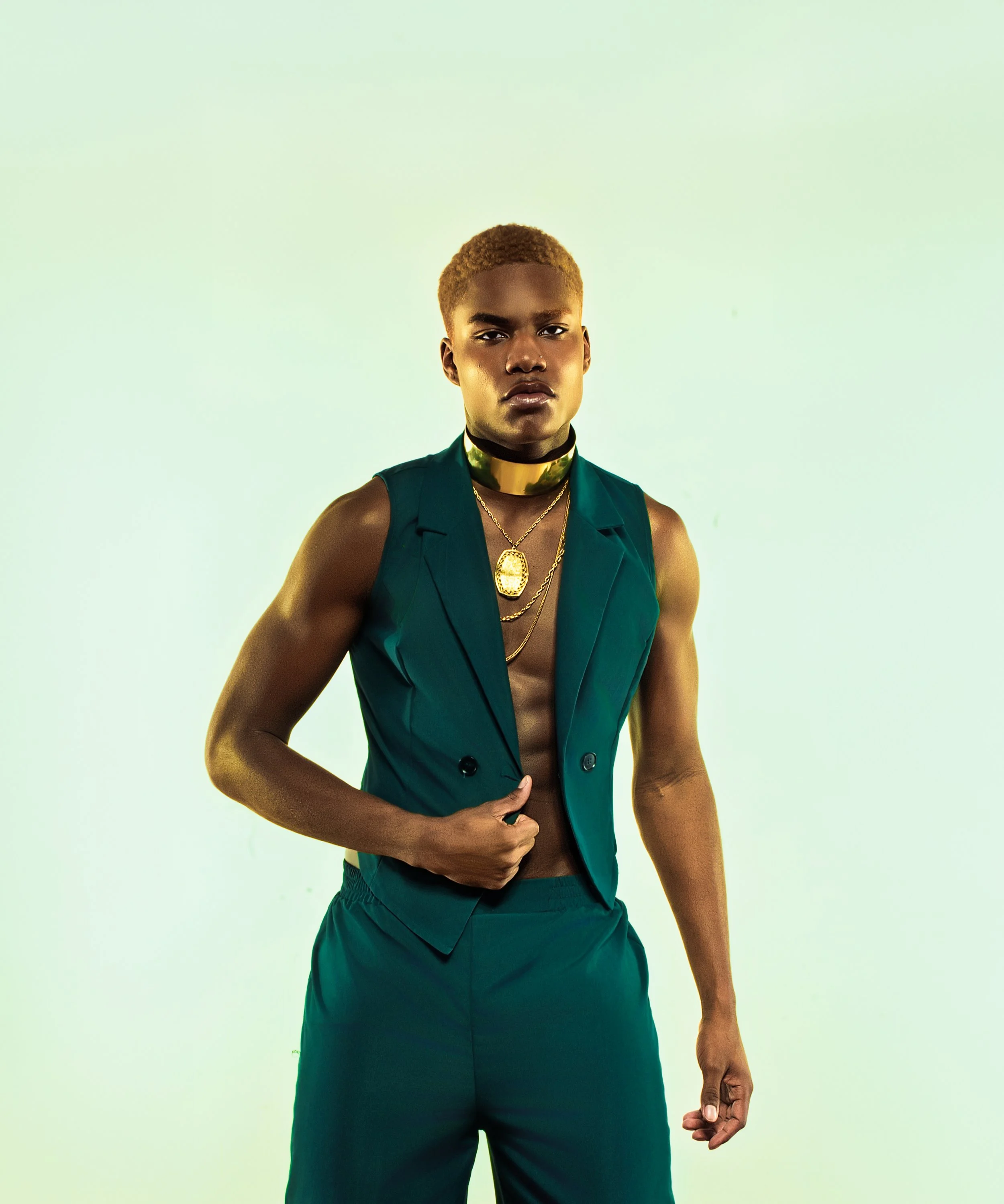 Person with short, light brown hair, wearing a teal sleeveless blazer, gold necklaces, and a shiny gold choker, standing against a light green background.