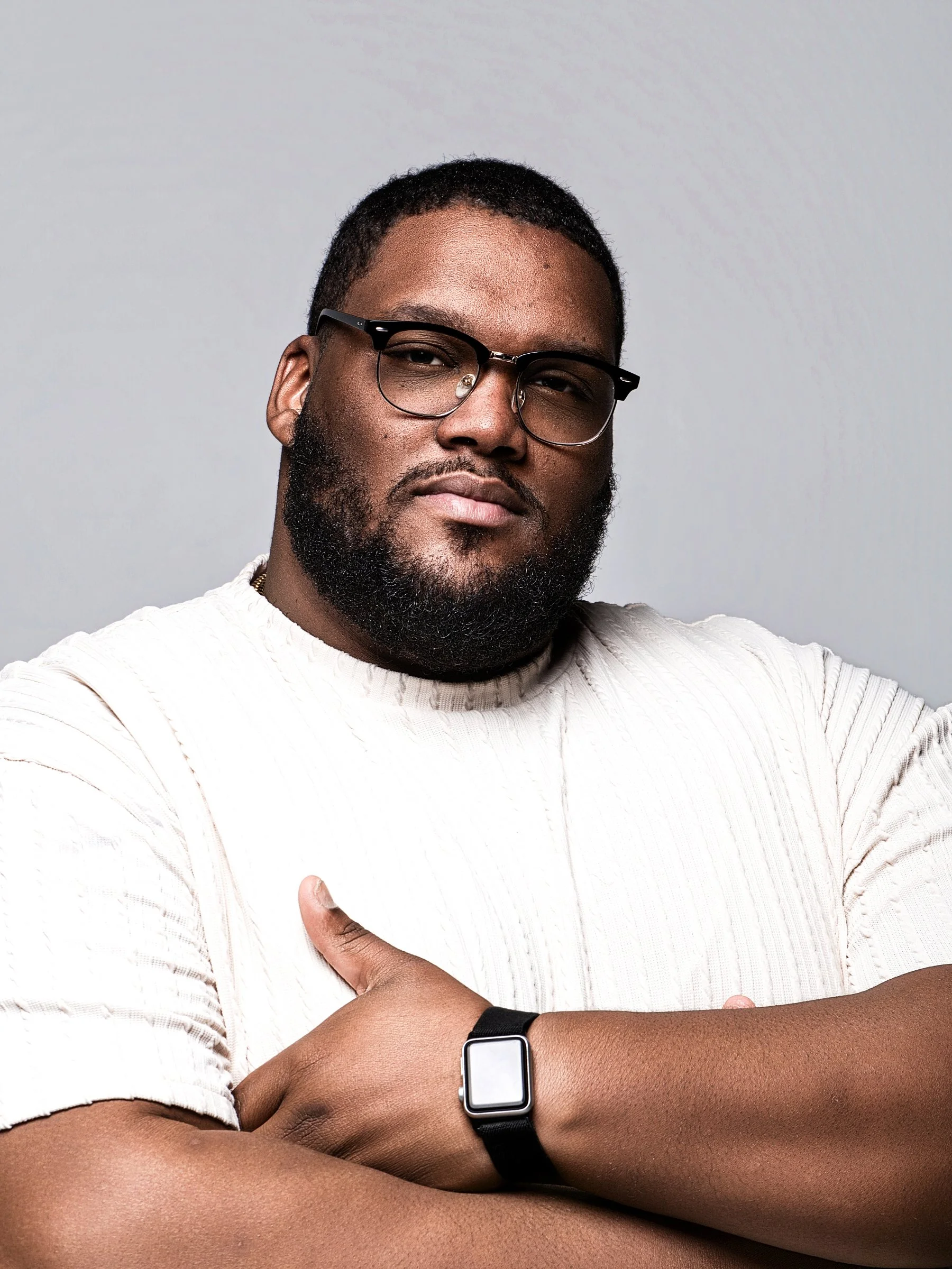 A Black man with glasses and a beard wearing a white sweater, with his arms crossed and a smartwatch on his wrist, against a plain gray background. Derick Jerome or Derick Todd. MUSEDPhoto