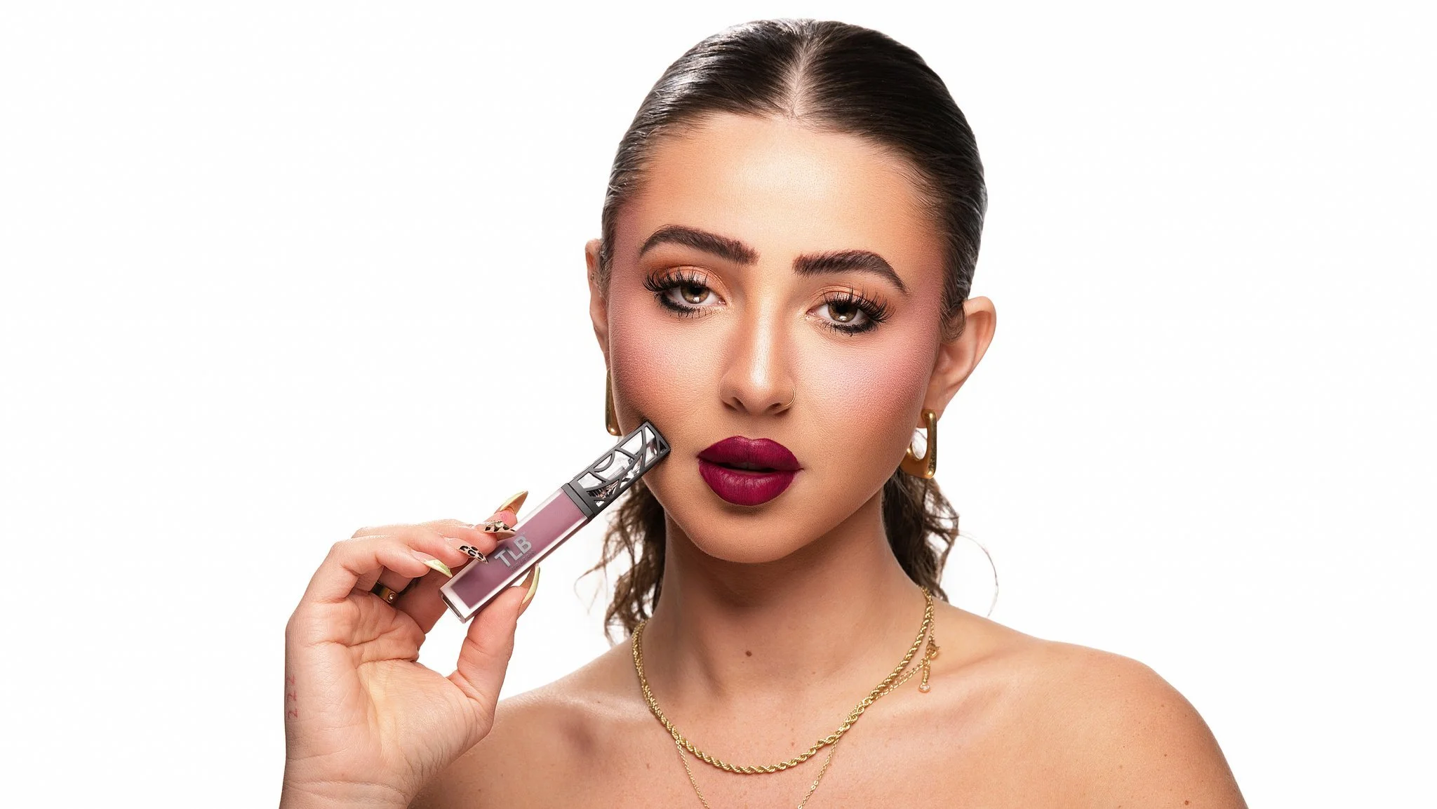 A woman with styled brown hair and makeup applying lipstick with a tube labeled TLB. She wears gold jewelry and poses against a white background.