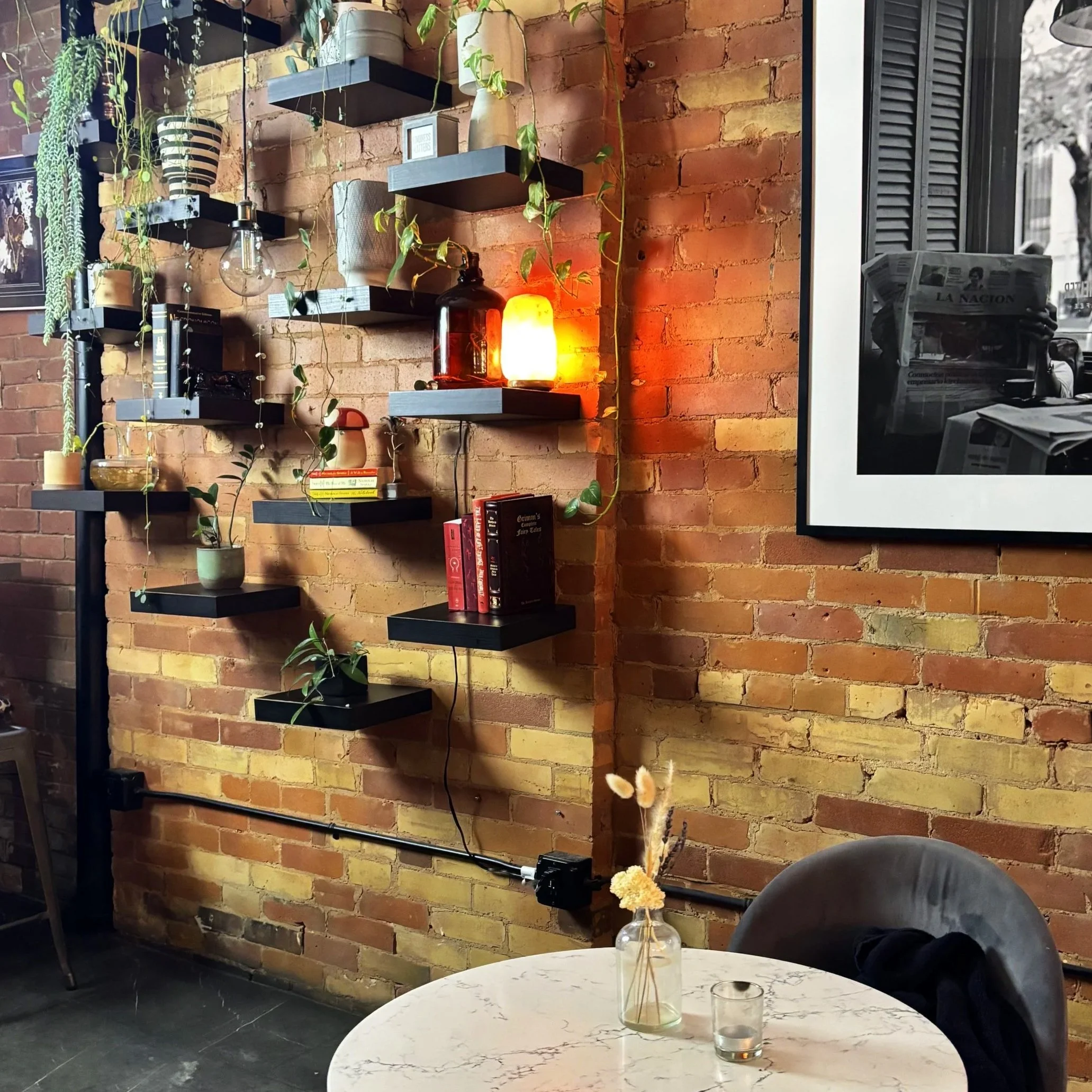 A brick wall with black floating shelves holding plants and small decorative items. A table with a flower vase and glasses is in the foreground, and framed black and white photos are on the wall to the right.