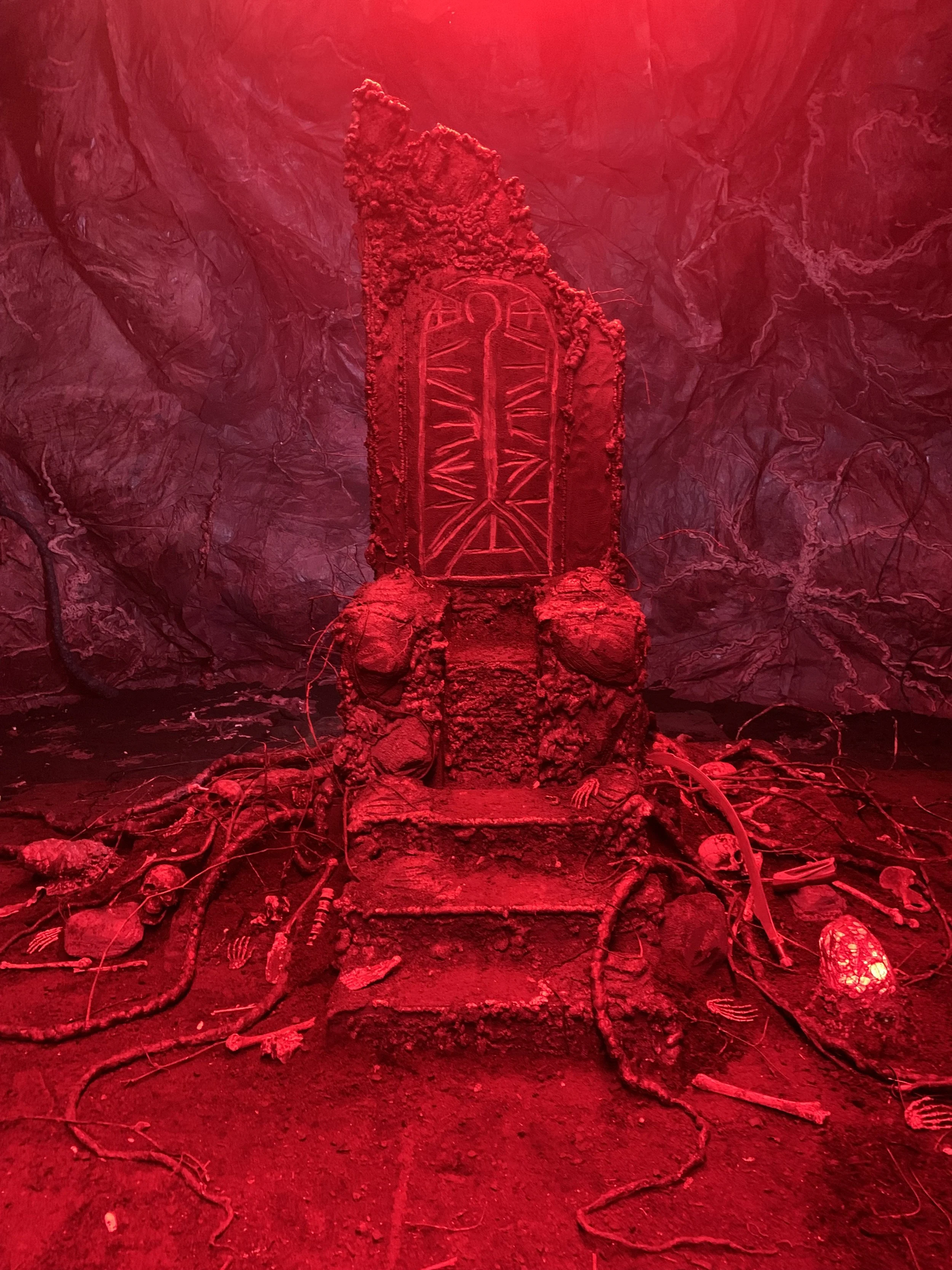 A metallic object resembling a tombstone or memorial surrounded by burned and damaged wires, with a red light shining overhead and a dark, textured background.