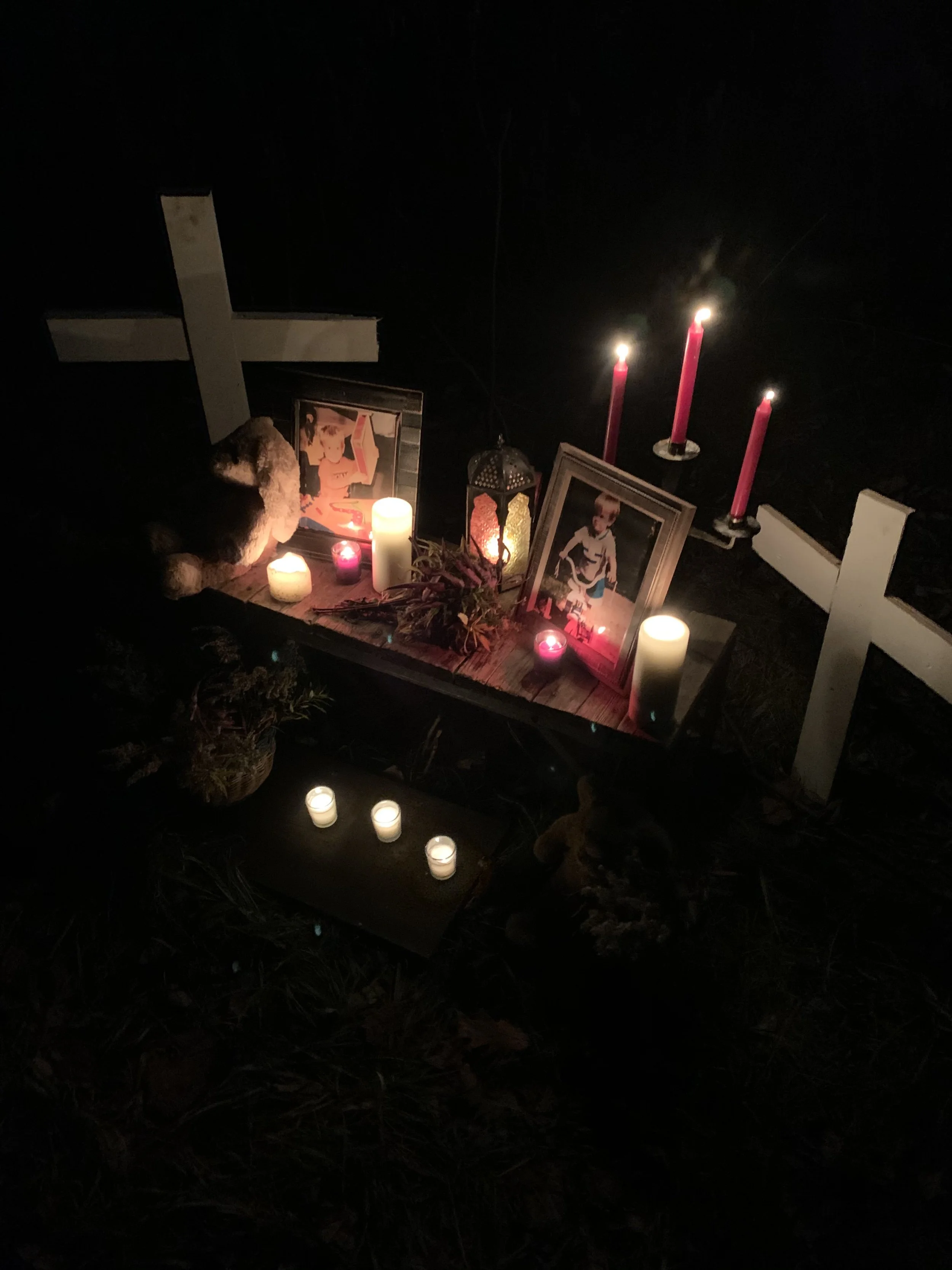 Memorial altar with candles, framed photographs of children, a white cross, pink candles, and decorative items, set outdoors at night.
