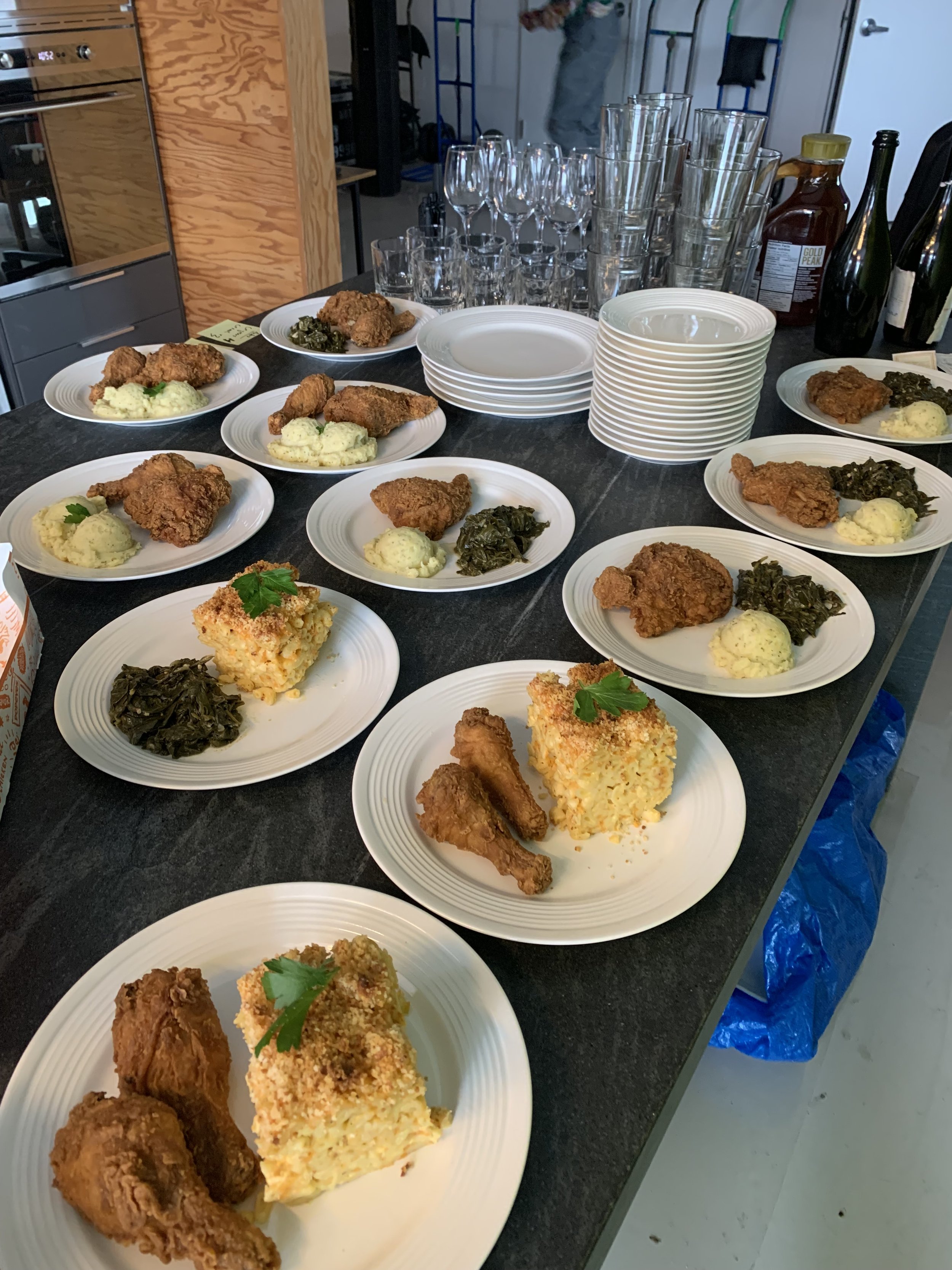 Plates with fried chicken, mashed potatoes, greens, and casserole on a table for a meal. There are empty glasses and stacks of plates nearby.