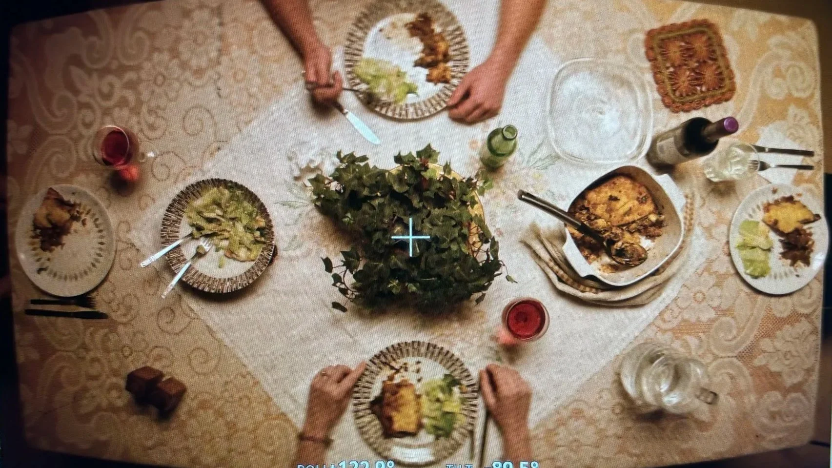 Top-down view of a family dinner table with plates of food, drinks, and a centerpiece, showing a person serving food on a decorated table with a beige tablecloth.