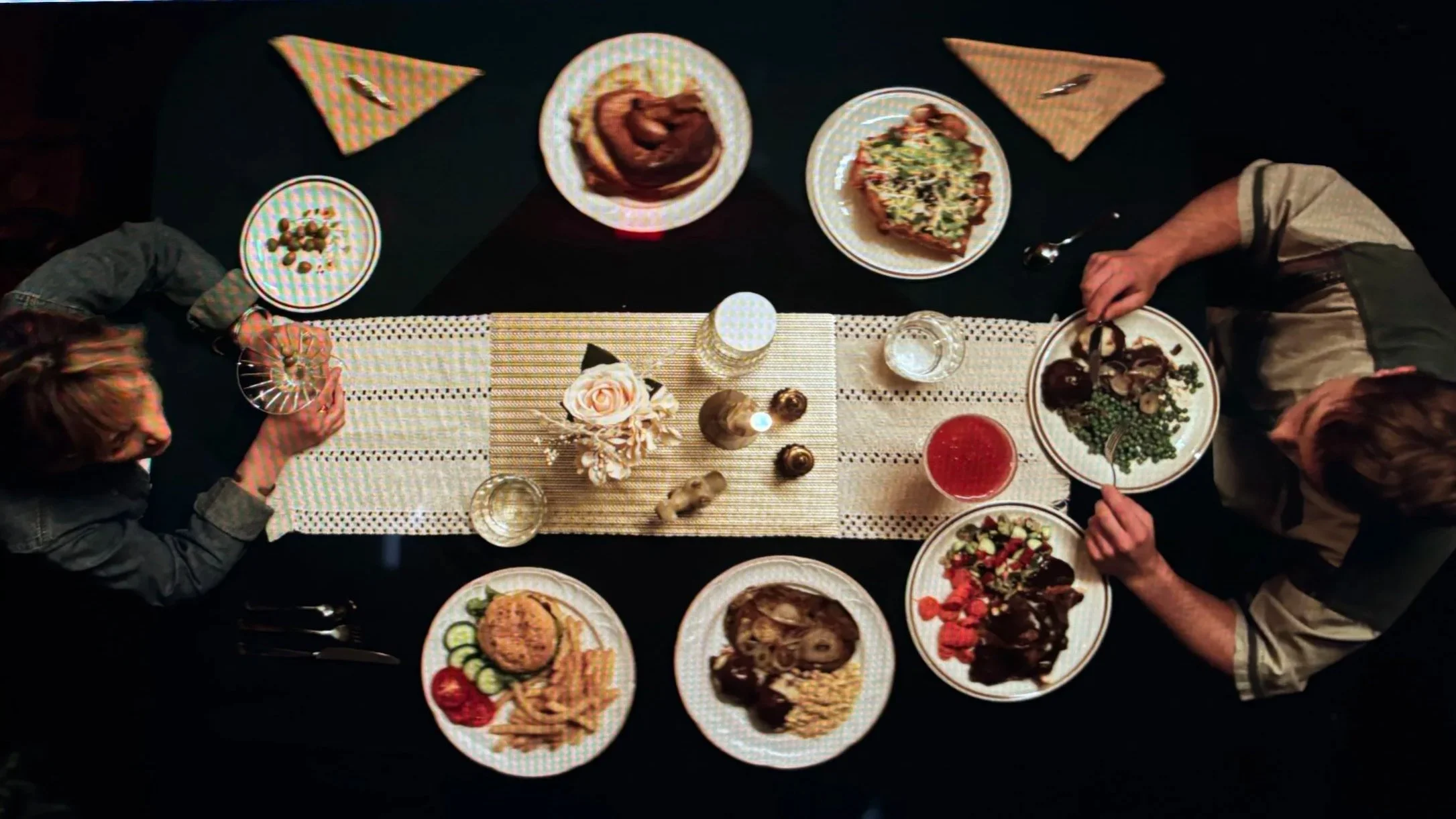Overhead view of a dinner table with various plates of food, drinks, and decorative items, surrounded by four people.