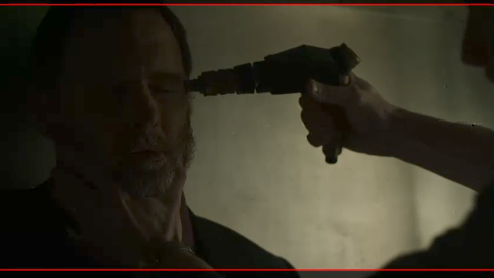 A person is holding a gun to another person's head in a dark setting.