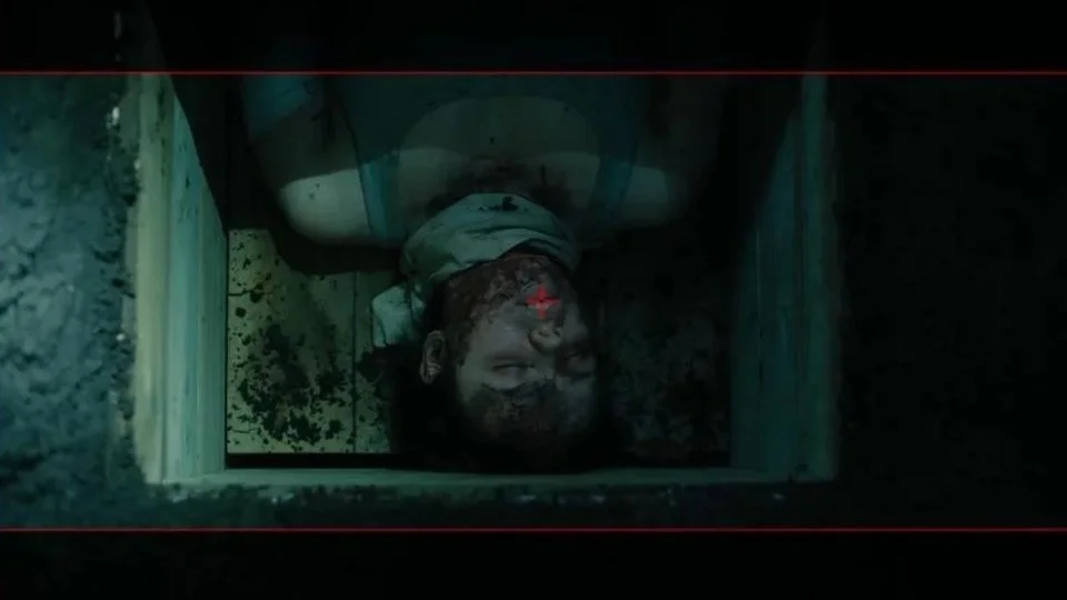 A person lying inside a narrow, dark wooden box or compartment, with their eyes closed and a bloody injury on their face, illuminated by a red laser or light beam. The scene appears intense and possibly related to an interrogation or horror setting.
