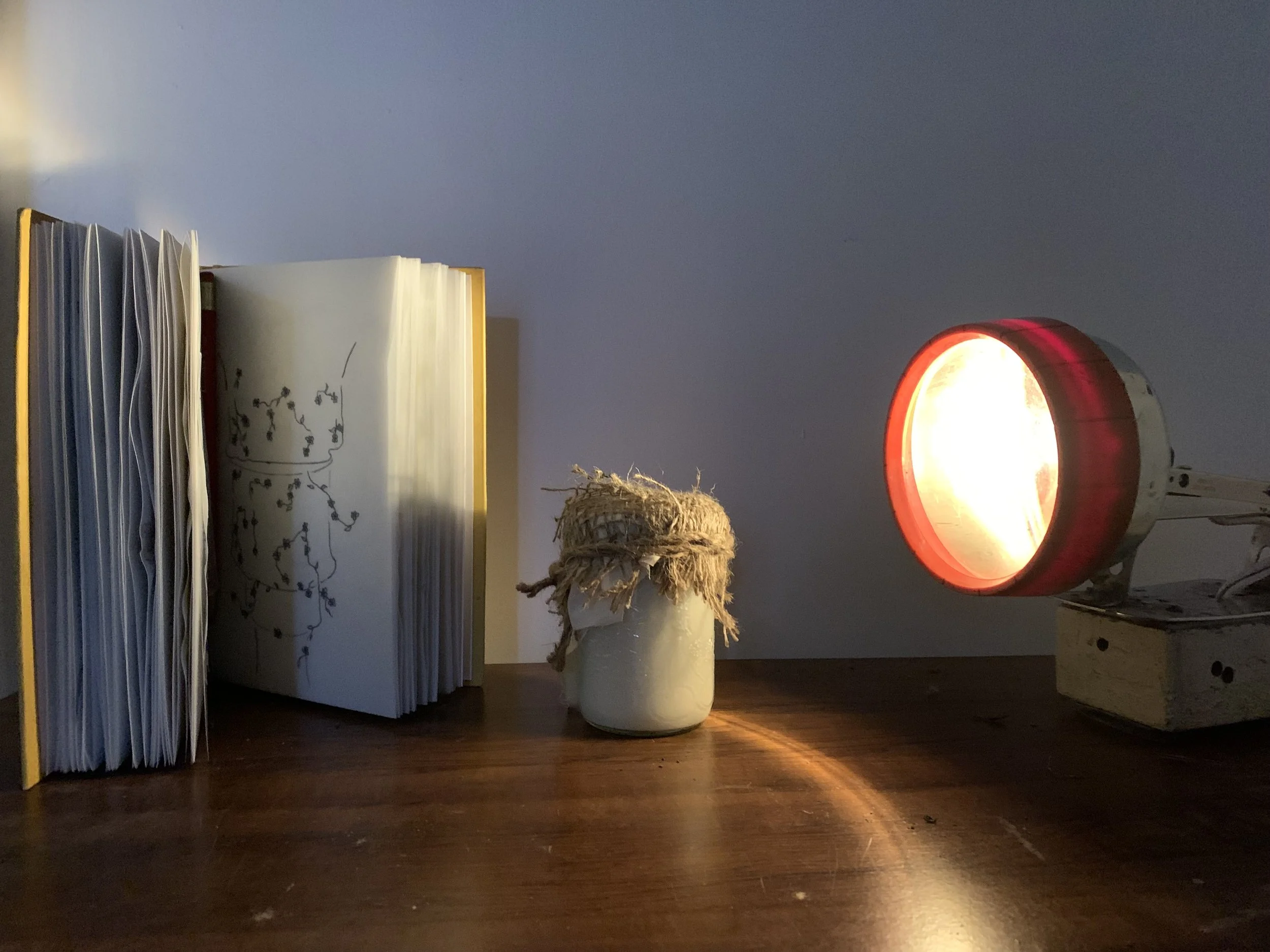 Beside a large lit orange lamp, there are several shelves with neatly organized notebooks or files on a wooden surface, a paper with a printed diagram or sketch, and some decorative items including a small plant and a woven ball.
