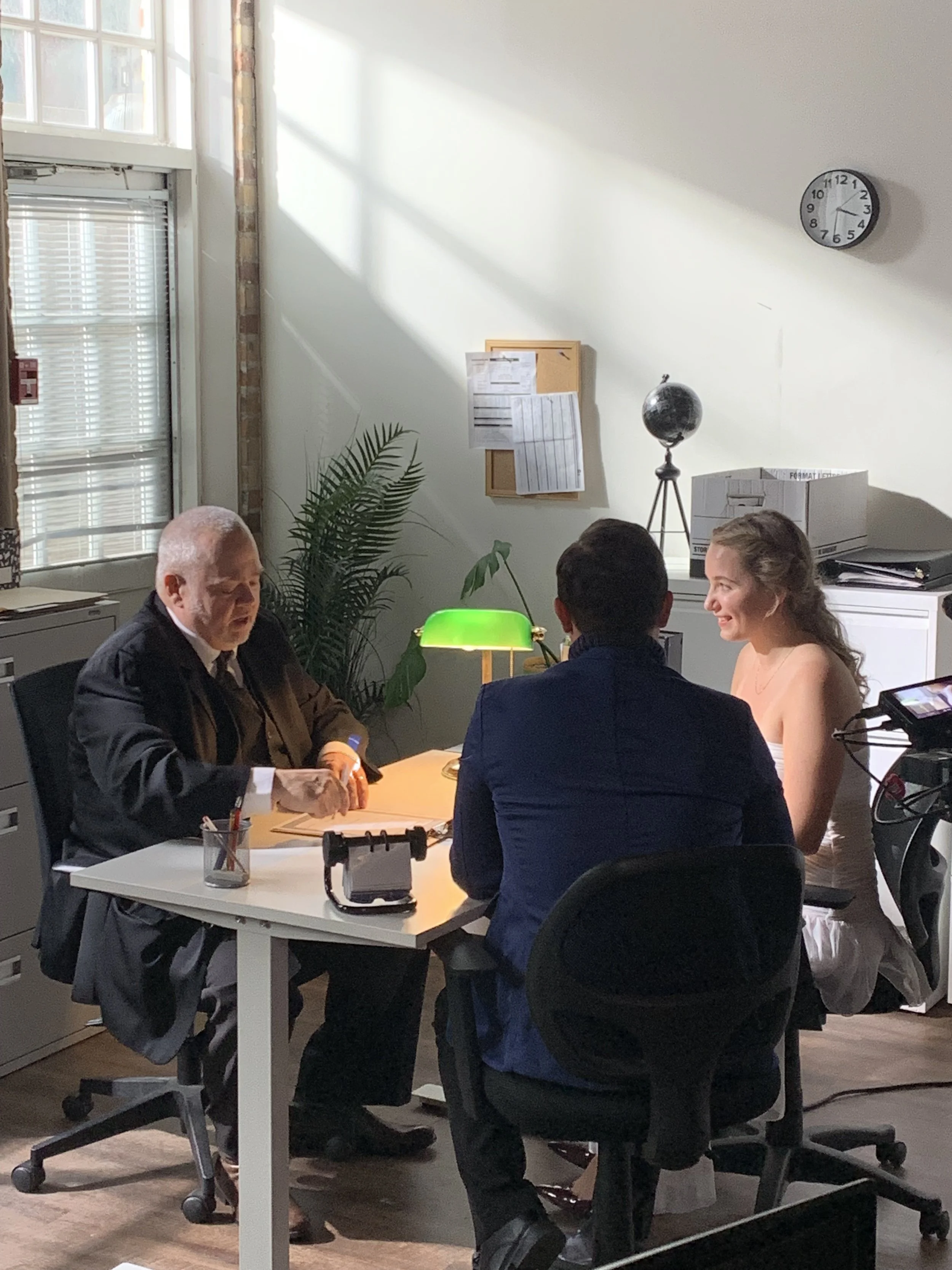People sitting around a white table in an office, with a light green desk lamp, a globe on a stand, a corkboard with papers, and a window with blinds in the background.
