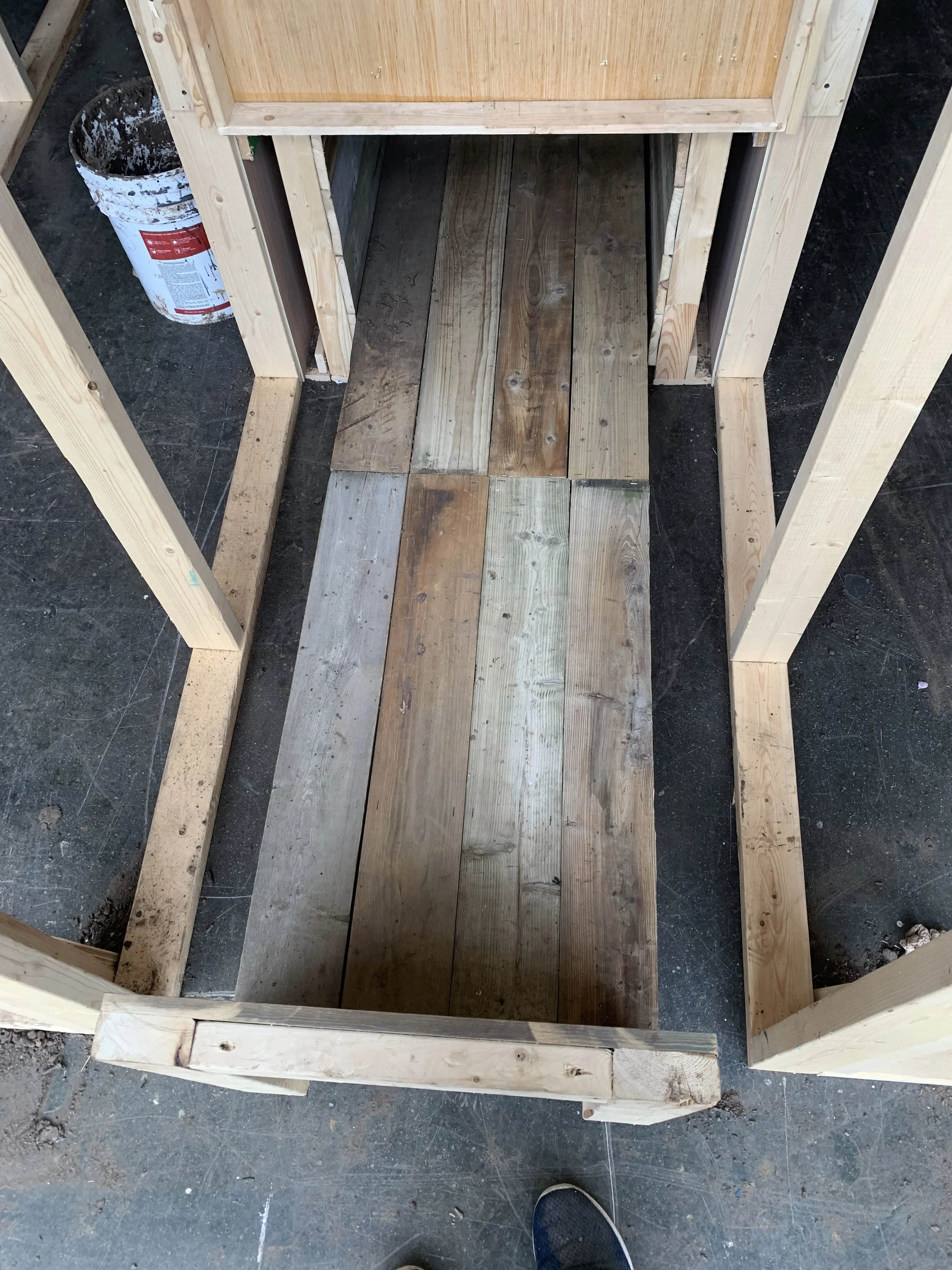 Wooden frame of a structure under construction with a wooden floor, a bucket of paint or stain to the side, and a person's shoe visible at the bottom of the image.