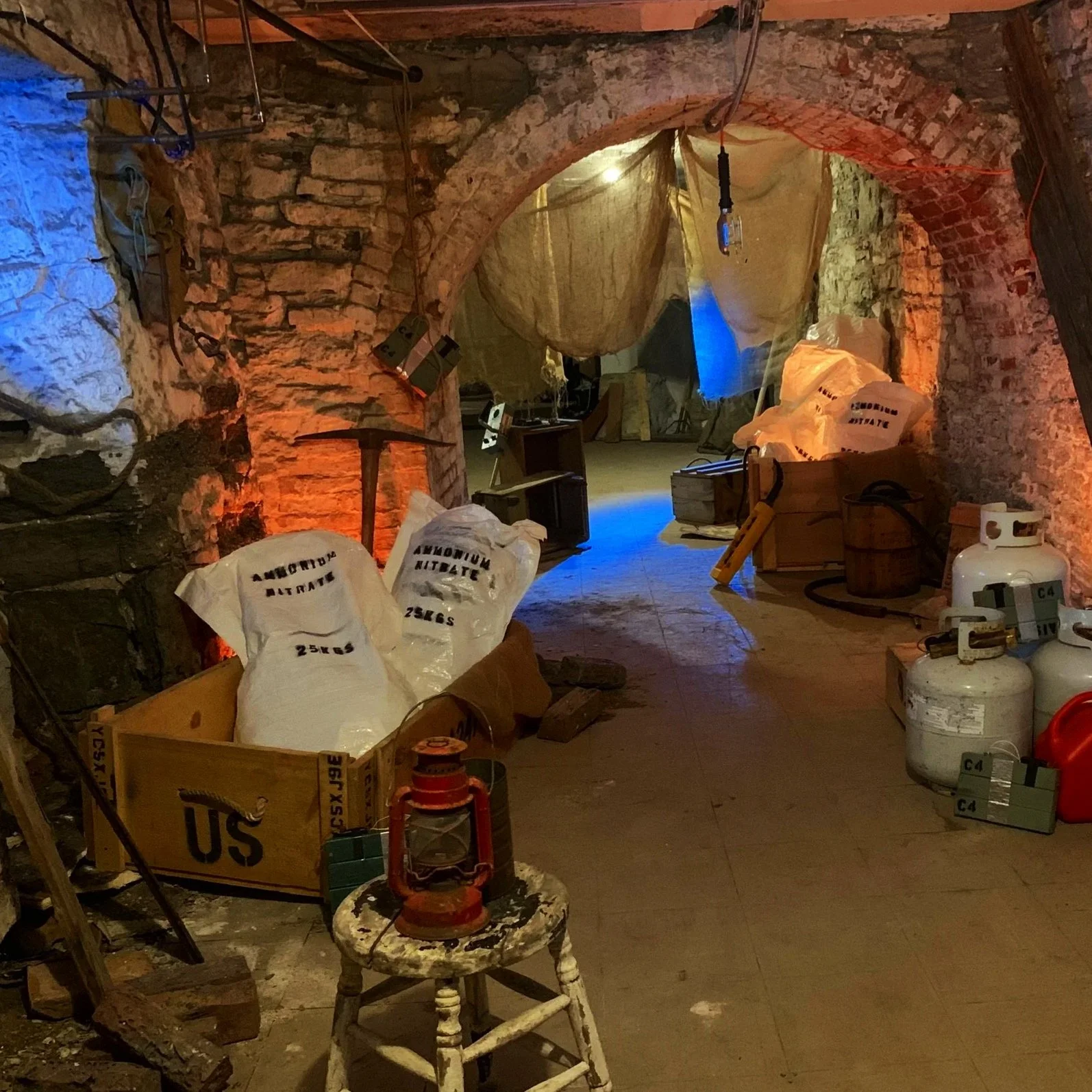 Underground storage space with brick archways, construction materials, tools, and equipment, including bags labeled 'Ammonium Nitrate,' a propane tank, a lantern, and a box marked 'US'.