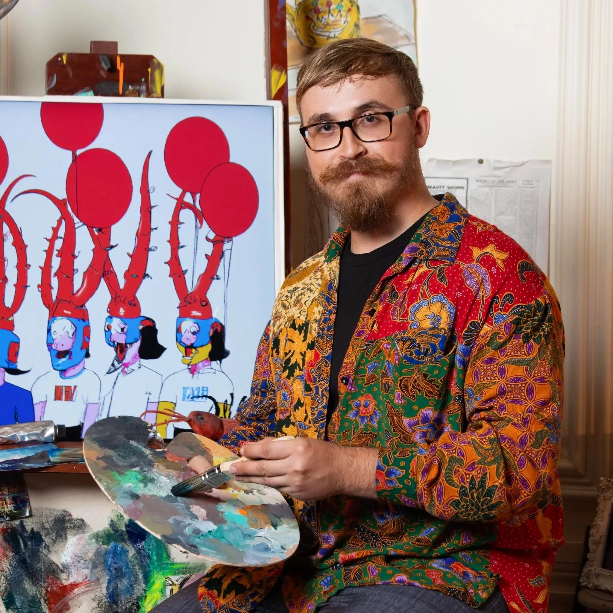 A man with glasses and a beard, wearing a colorful patterned shirt, is holding a paint palette and standing next to a computer screen displaying cartoon characters. The background shows a white wall with a painting and some papers.