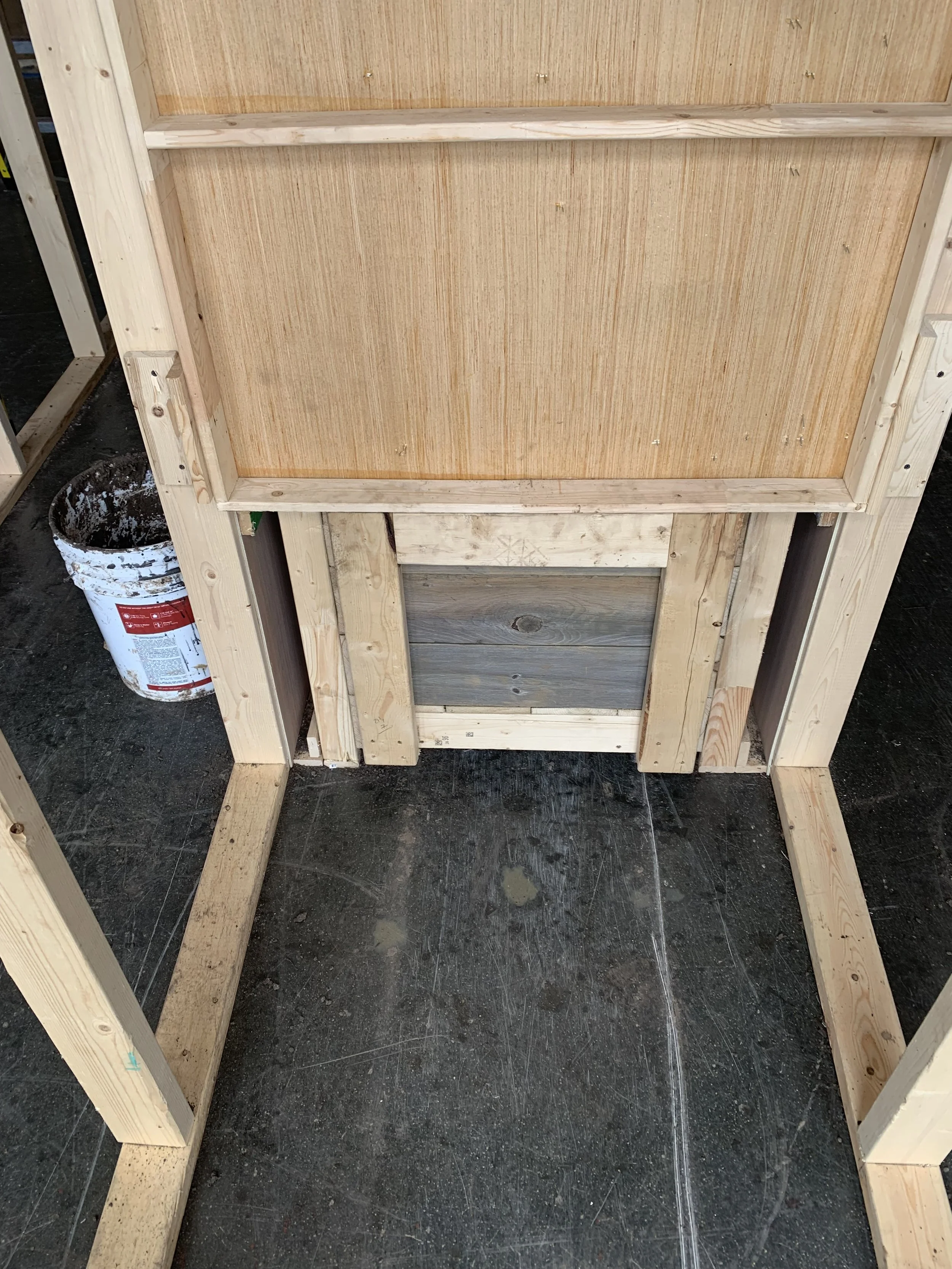 The image shows a wooden frame for a piece of furniture, possibly a table or a counter, under construction. There is a bucket of paint or stain on the floor nearby, and the workspace appears to be in a workshop or construction area with a concrete or