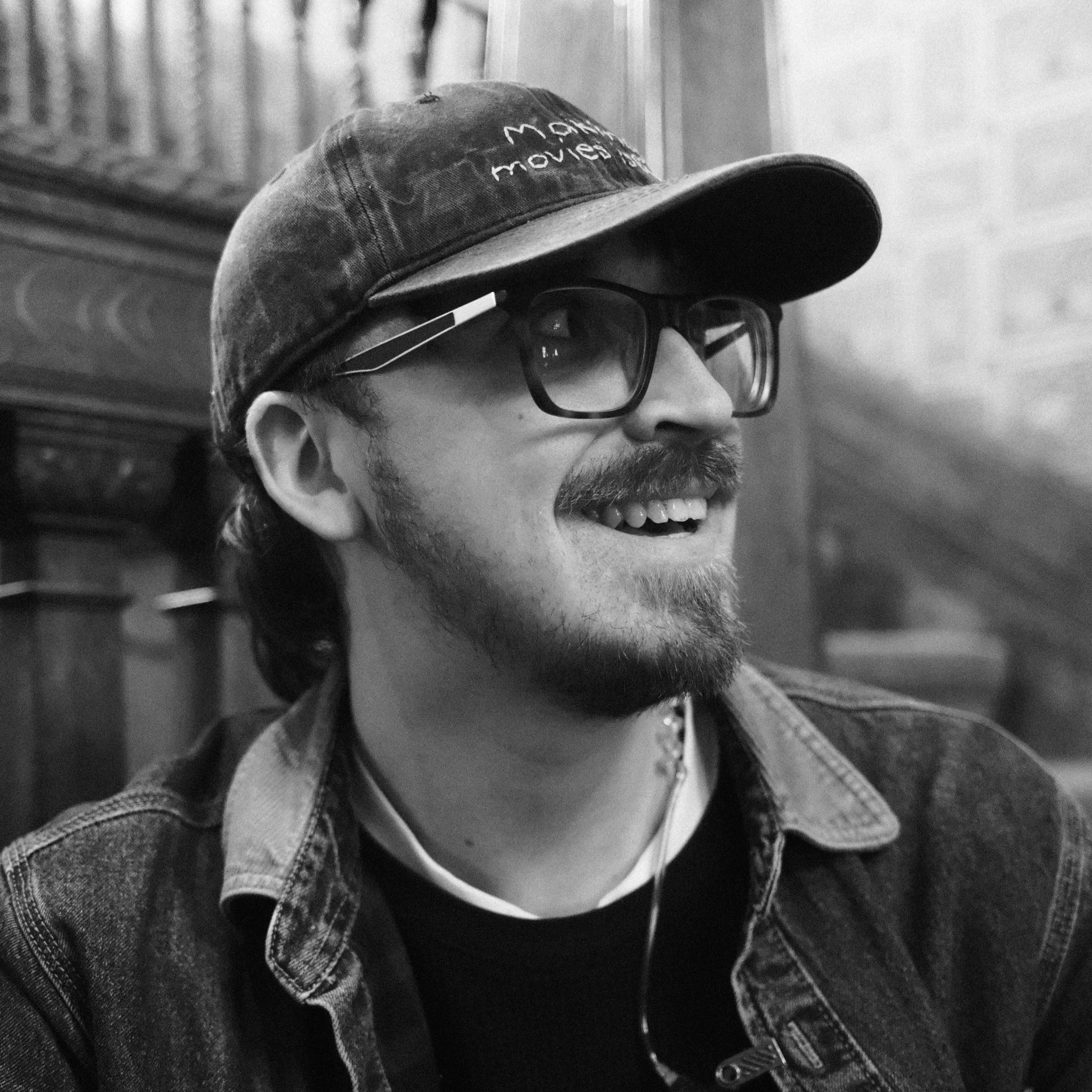 Close-up of a person wearing a baseball cap, sunglasses, and denim clothing, smiling and looking to the right.
